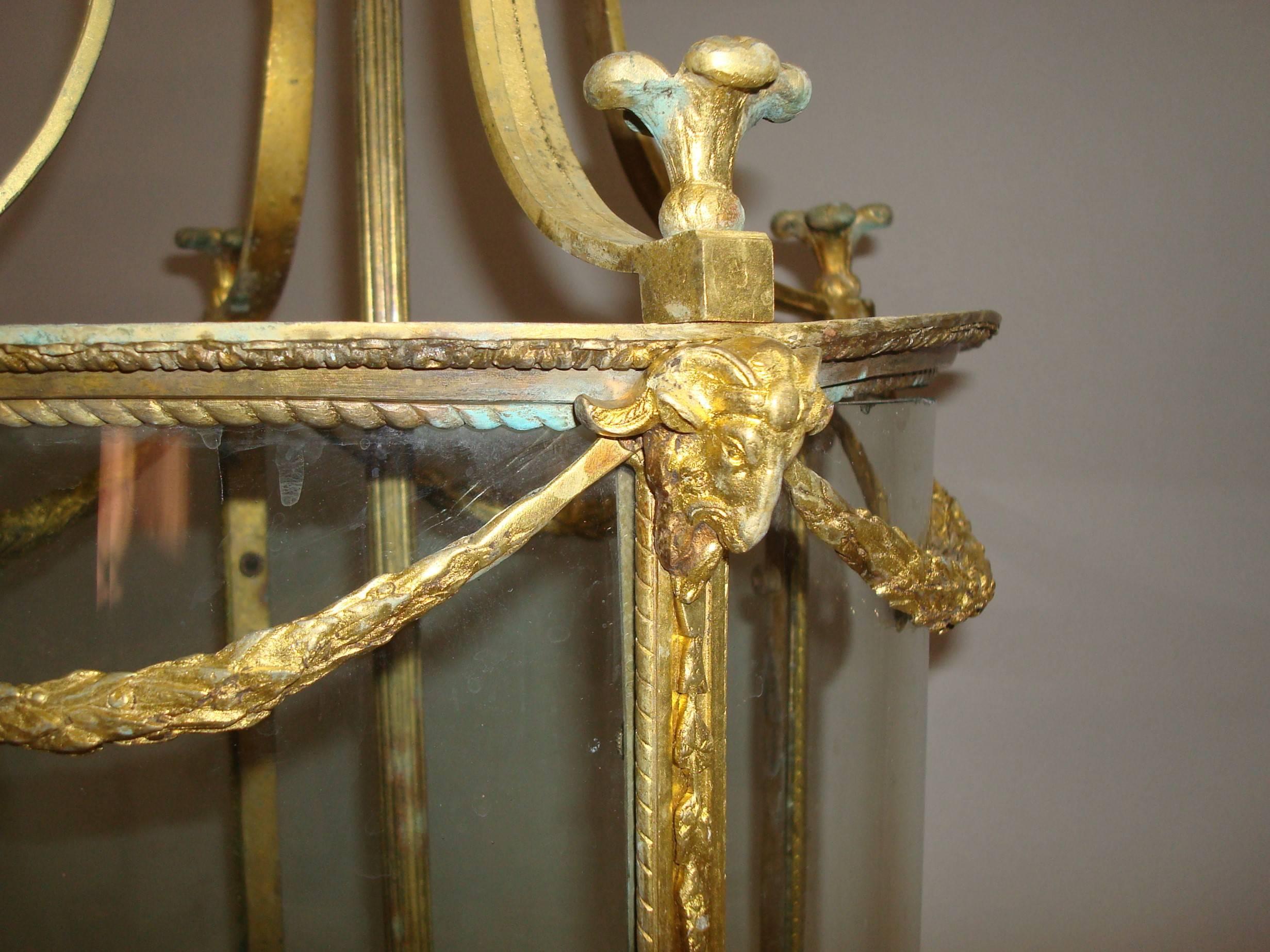 19th century gilt brass hall lantern of cylindrical form in the neo classical taste; the circular boss with hanging loop and four swept arms with palm finials and rams heads below joined by laurel leaf swags; the circular glass in a framework with