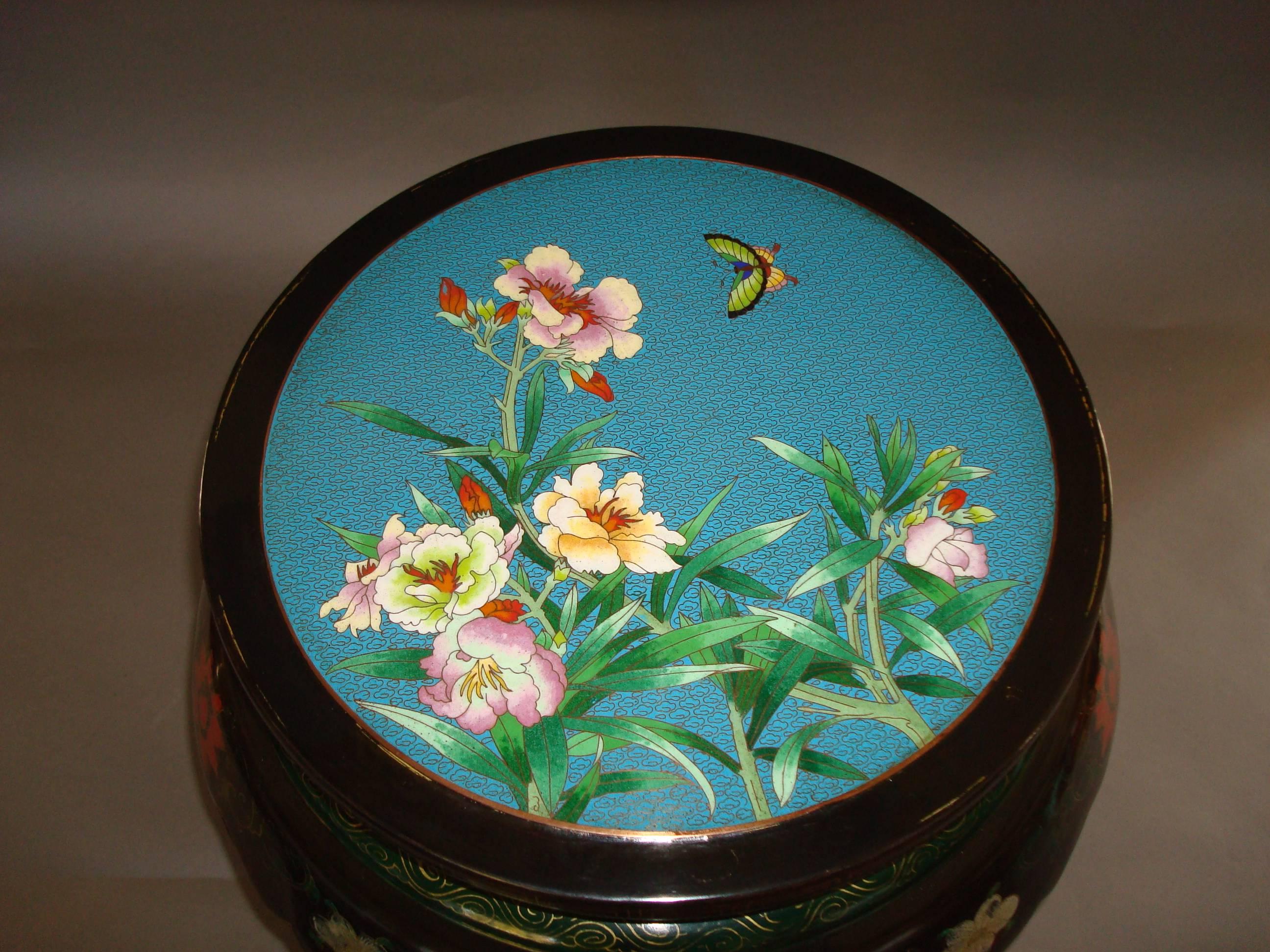 A pair of Chinese lacquered and cloisonné low tables; the circular tops inset with colourful cloisonne enamelled panels, one with a spray of flowers and a butterfly, the other with a vase of flowers and Chinese objects.  The moulded edge with gilt