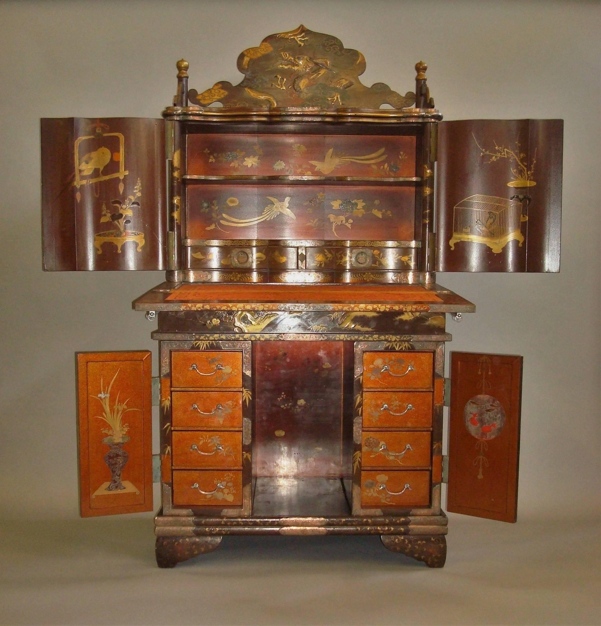 An excellent 19th century Japanese writing cabinet of unusual design, small proportions and profusely decorated; the shaped back with a dragon scene and gilt finials to the corners, above a pair of serpentine panelled doors depicting lions in a