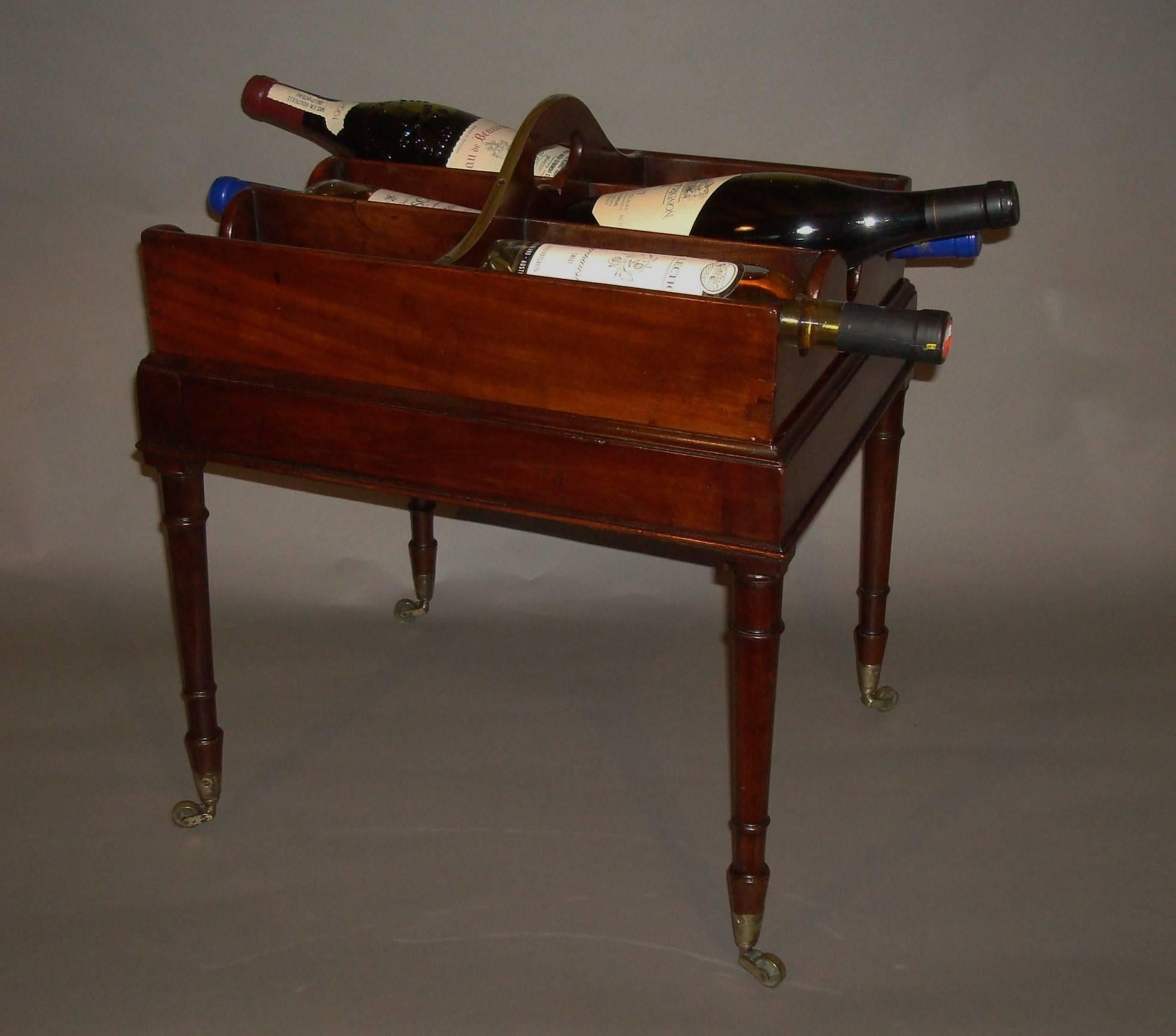 An unusual George III mahogany bottle carrier on stand; the removable bottle tray with a shaped central division and a pierced carrying handle finished with a brass moulded edge; having six divisions for wine bottles with shaped sides to house the