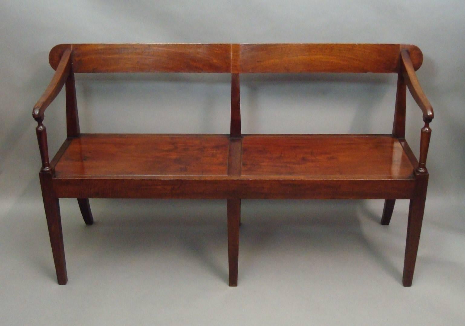 A good George III mahogany hall seat; the figured mahogany crest rail with a twin curve and rounded ends the swept arms, on slender turned supports, leading to the well figured double panel seat with a narrow reeded edge, set in a plain seat frame;