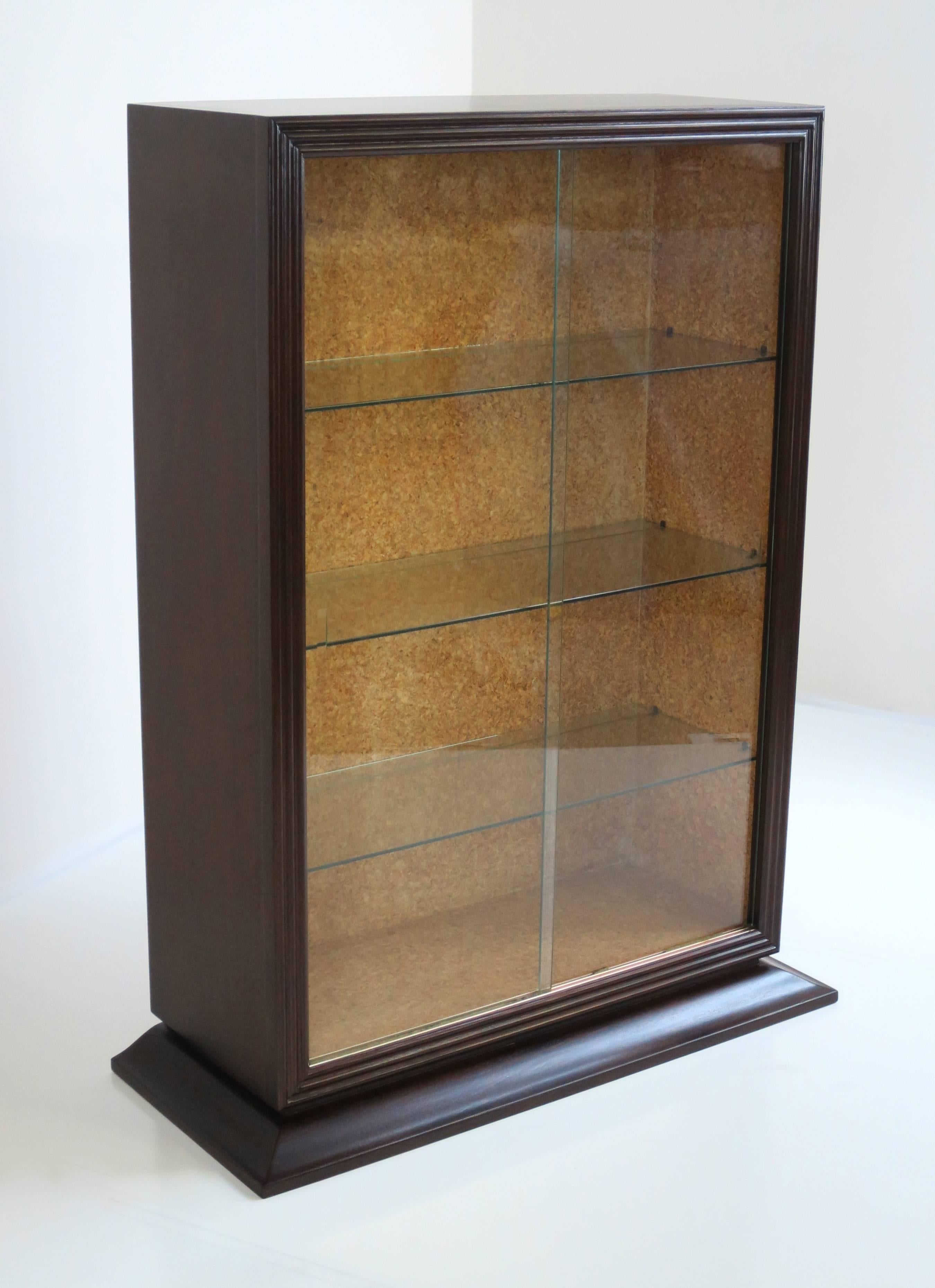 Paul Frankl for Brown Saltman Vitrine. Cork lined interior with glass shelves and sliding glass doors. Mahogany construction with flared and stylized base. By removing glass and replacing shelves with wood, this piece is ideal for a small bookcase