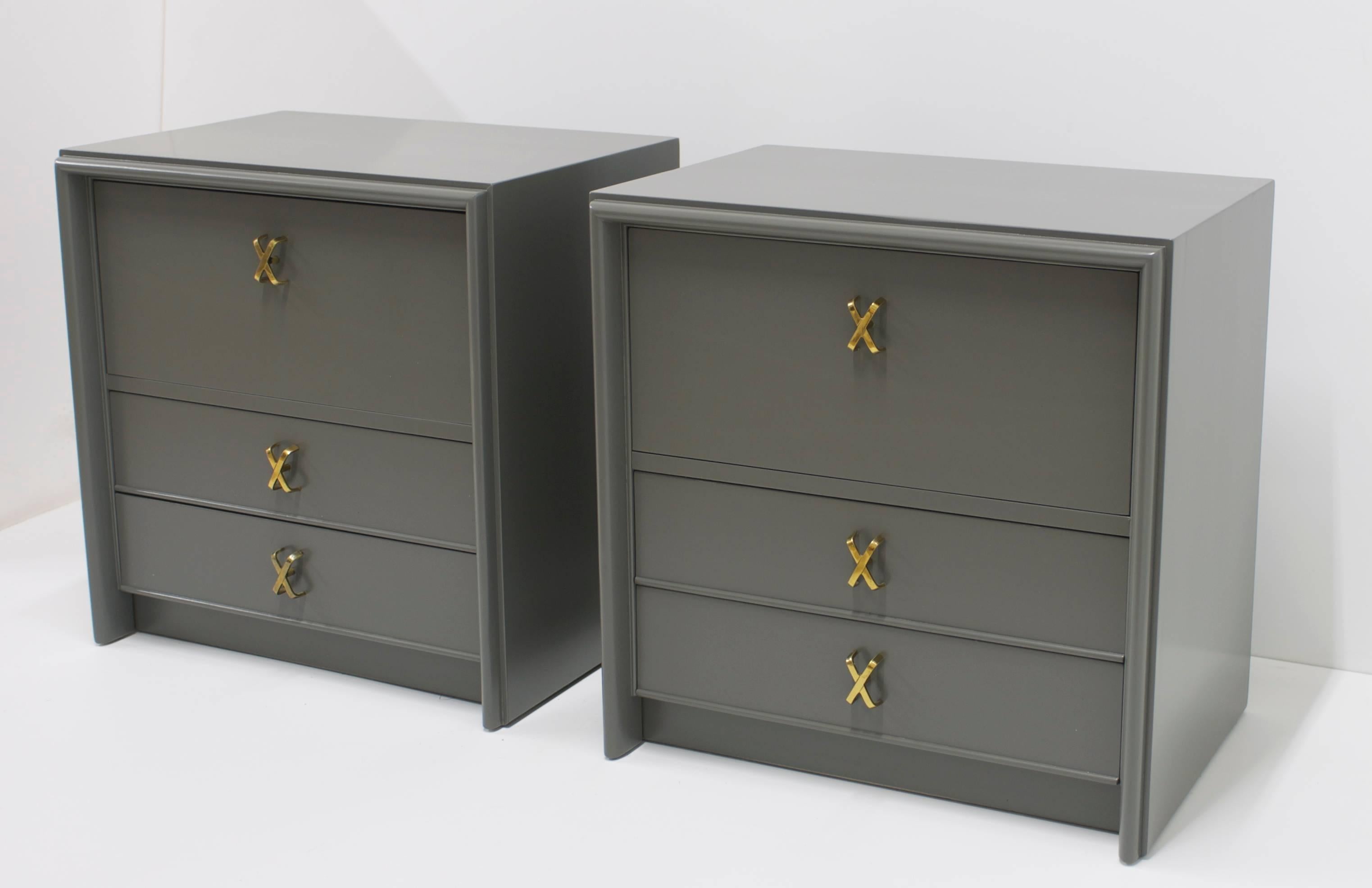 Designed by Paul Frankl for the Johnson Furniture Company, these pair of bedside tables and been refinished in a satin gray. The cabinets retain their original brass X-pulls. The top doors of each cabinet pull down to create a little shelf when