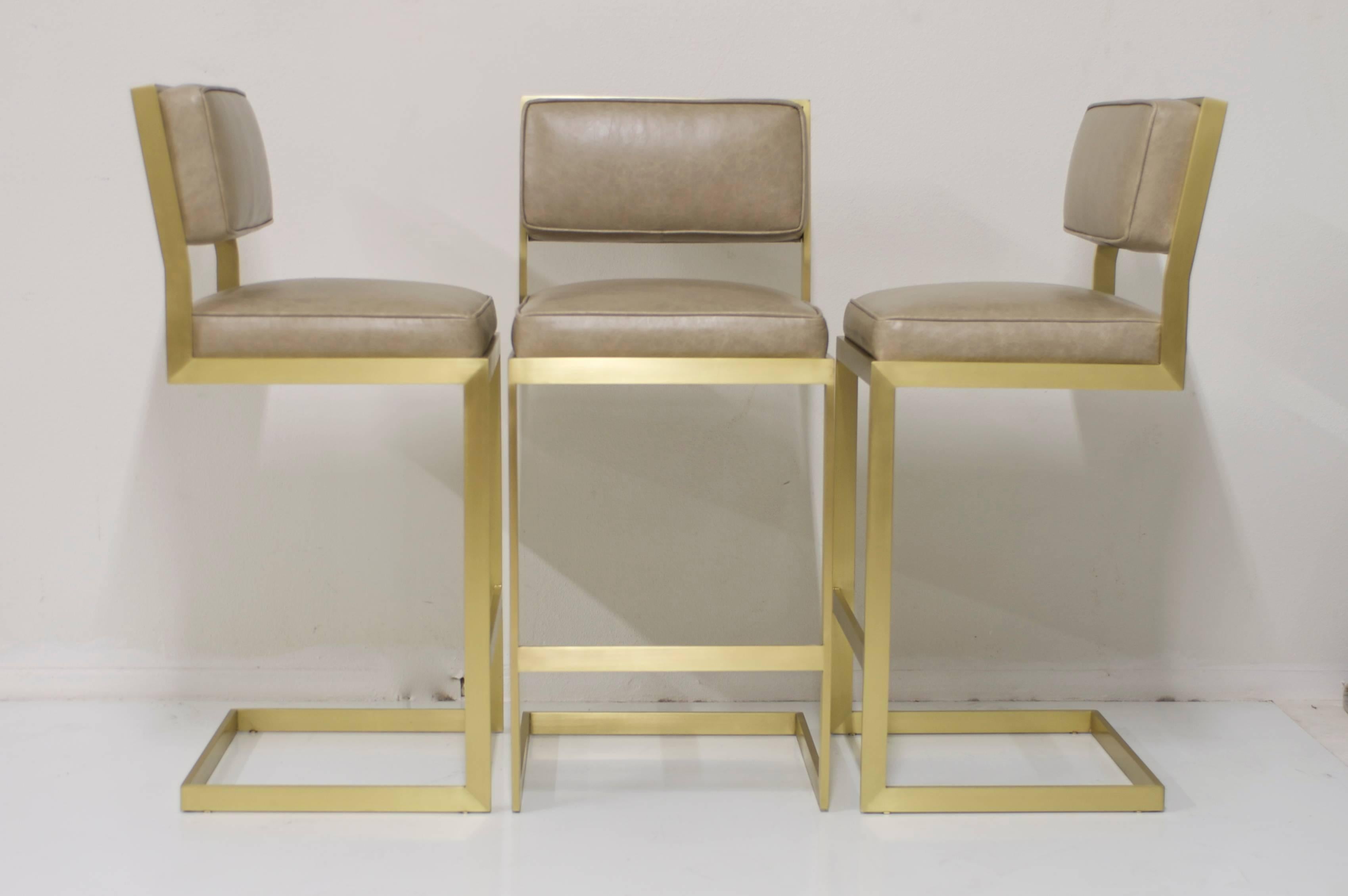 In the style of Milo Baughman this set of three bar stools have been plated in a beautiful brushed matte brass finish. They are a nice modern rectangular design made with flat bar steel and have a footrest. The seat and back are upholstered in a