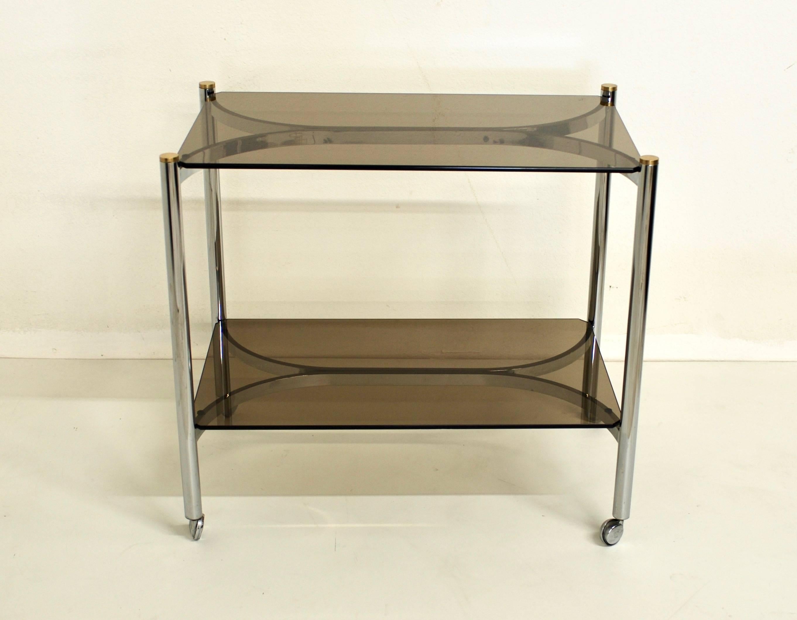 A mixed metal chrome 1970s era bar cart with brass caps and bronze colored glass. It rests on metal casters. The design of the cart has nice clean lines.