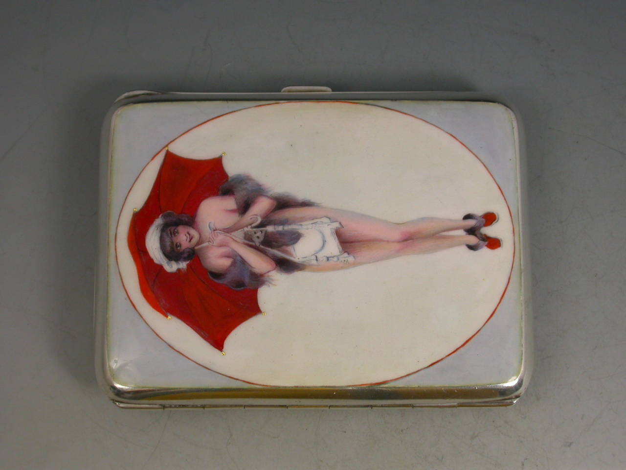 A fine quality early 20th century silver and enamel cigarette case of rounded rectangular form with sprung opening mechanism, the cover enameled with a young lady wearing just a mink stole and red shoes, holding a white handbag and a red umbrella.