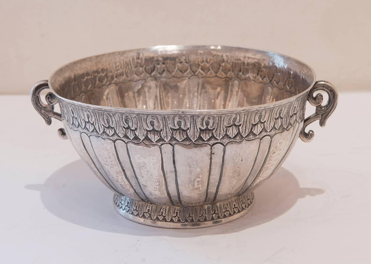 Circular, on circular foot, with lobed lower body, the slightly flared rim engraved with repetitive leaf motif, with two foliate scroll handles. The inside base of the bowl is engraved with three roses encircled by a wreath. Marker's marks are on