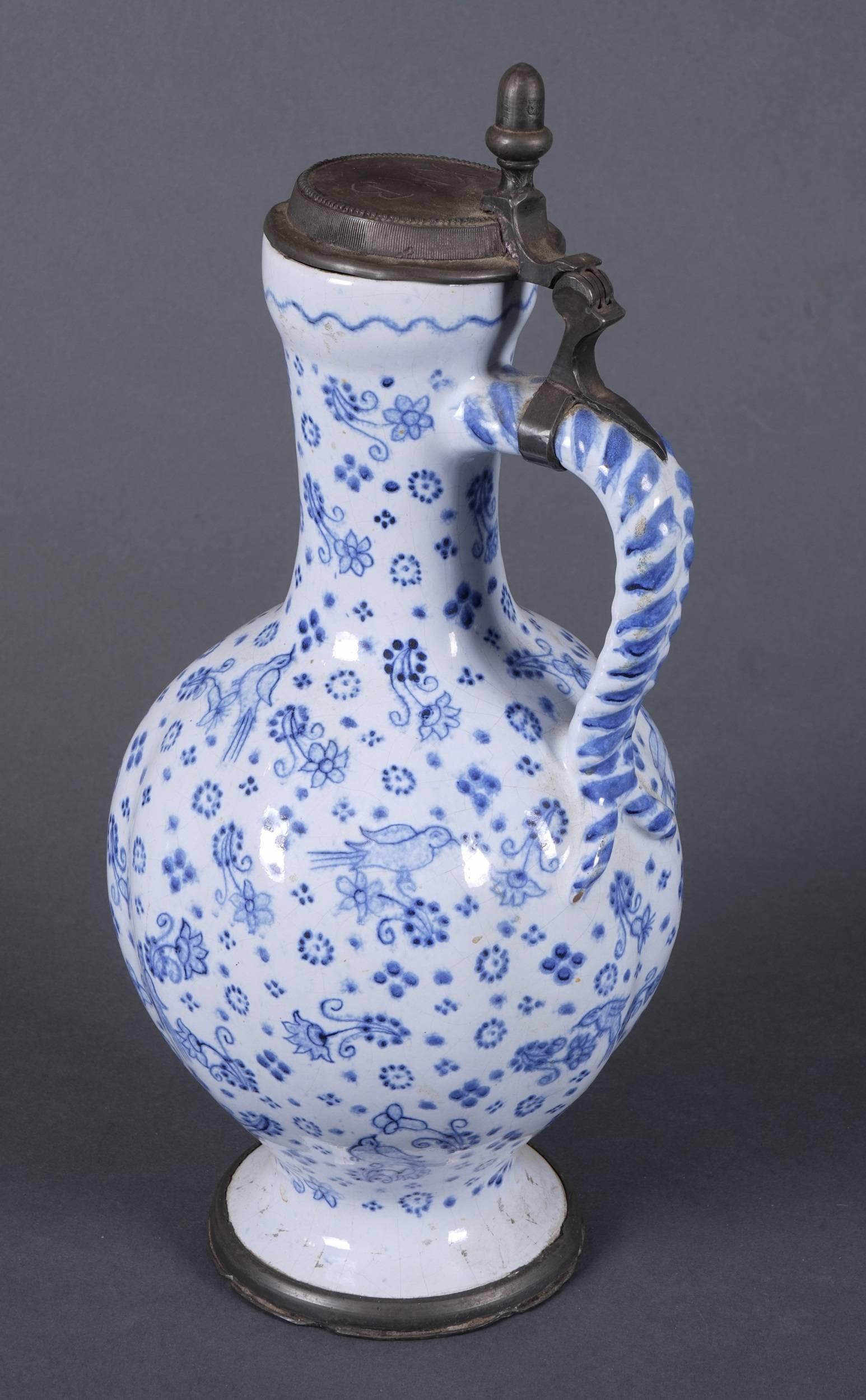 A blue and white pewter-mounted Enghalskrug or ceramic jug, the body decorated with a design of birds and flowers. The hinged cover is embellished with an acorn knop and inscribed 'KMB.'
   