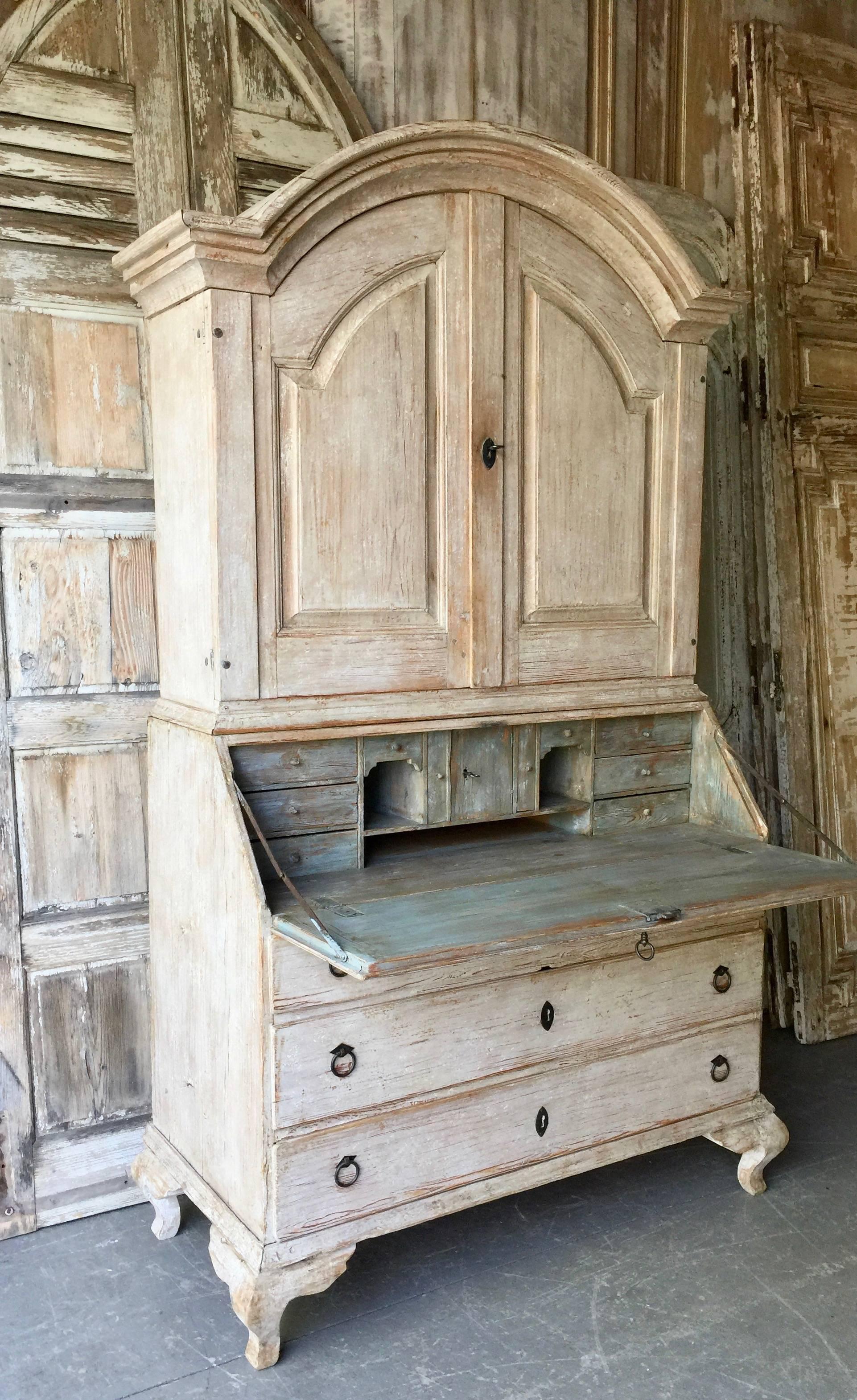 18th century period Swedish Rococo secretaire cabinet scraped to original grey or cream paint with arched pediment over raised panel doors with two interior shelves and notched spoon shelf. Fall front desk with multiple small compartments in worn