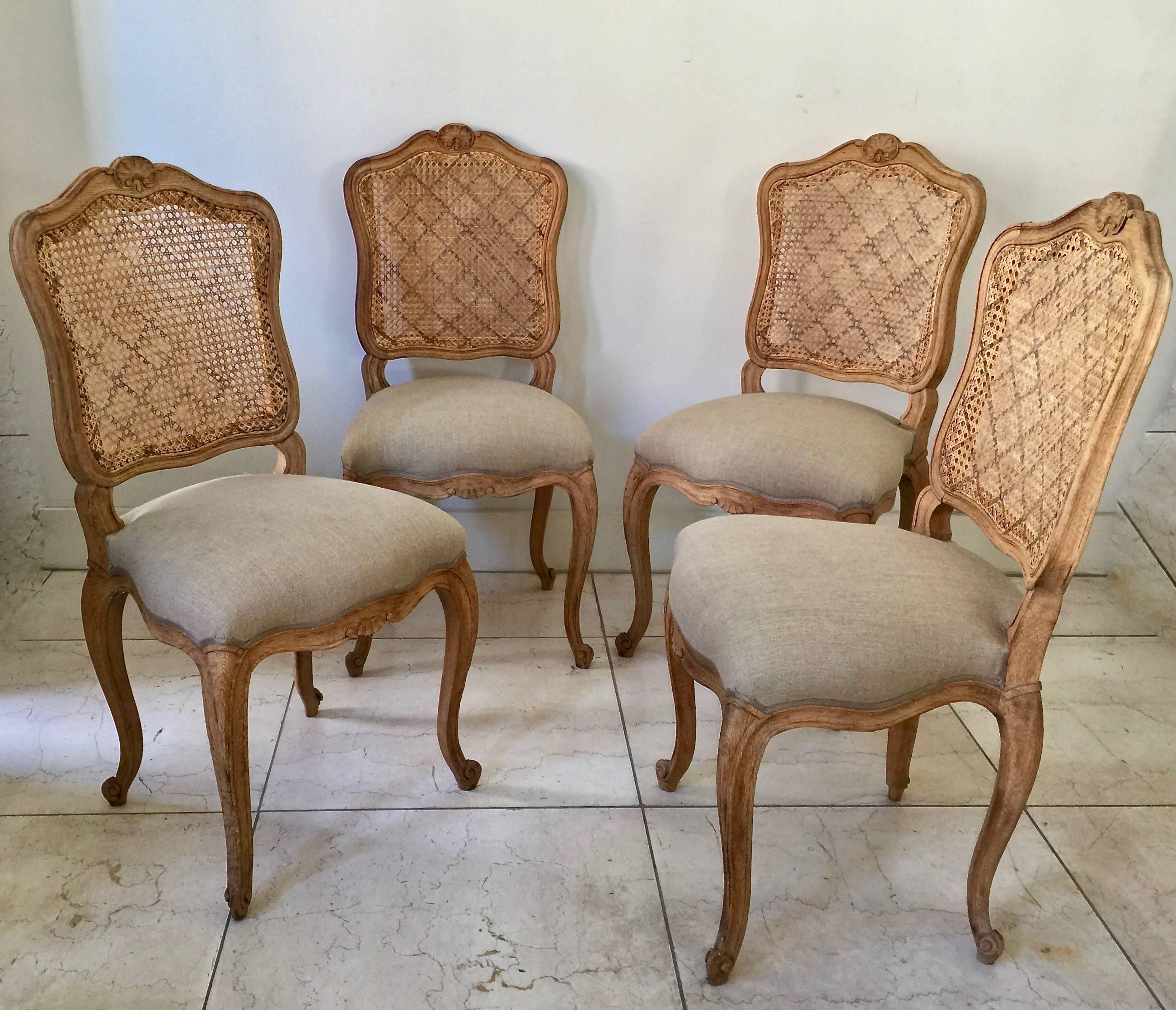 Set of four in natural oak French chairs Louis XV style with flat back called “la Reine” is caned and scalloped frieze carved and raised on cabriole legs. Upholstered in natural linen with classical decorative gimp trim. Very elegant as well as