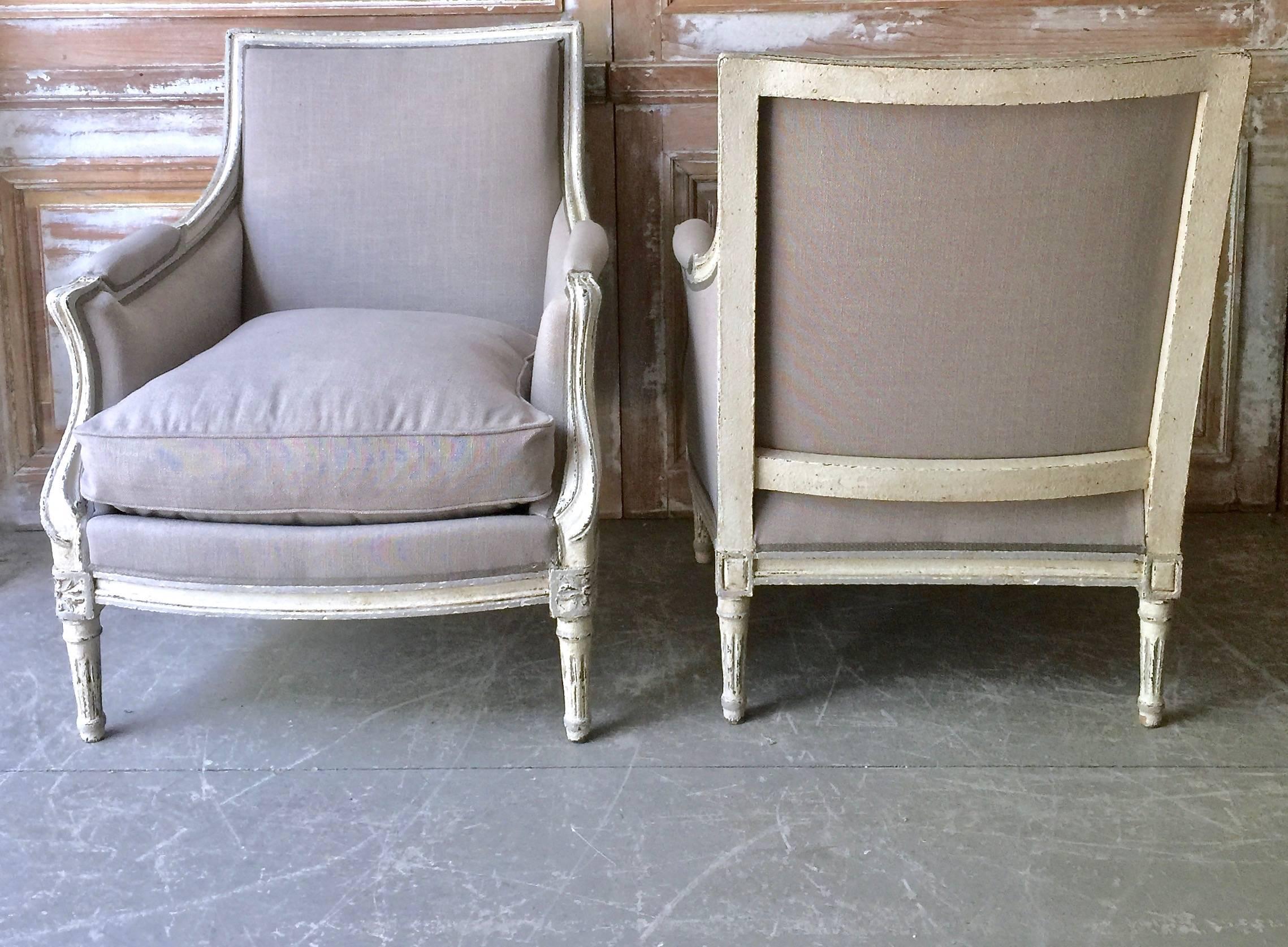 A pair of French painted Bergères in Louis XVI taste, with generous seats. The wood frames are in lovely blue-grey painted finish with richly carved frame, fluted leg and cubic blocks decorated with florets. Completely re-upholstered in linen