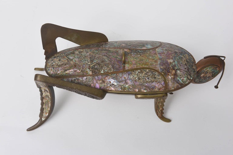 This whimsical and exotic lidded brass dish in the shape of a grasshopper is inlaid with copper and naturally iridescent abalone. The work is unsigned and unmarked but is in the style of Salvador Teran who worked in Los Castillo, Mexico in the