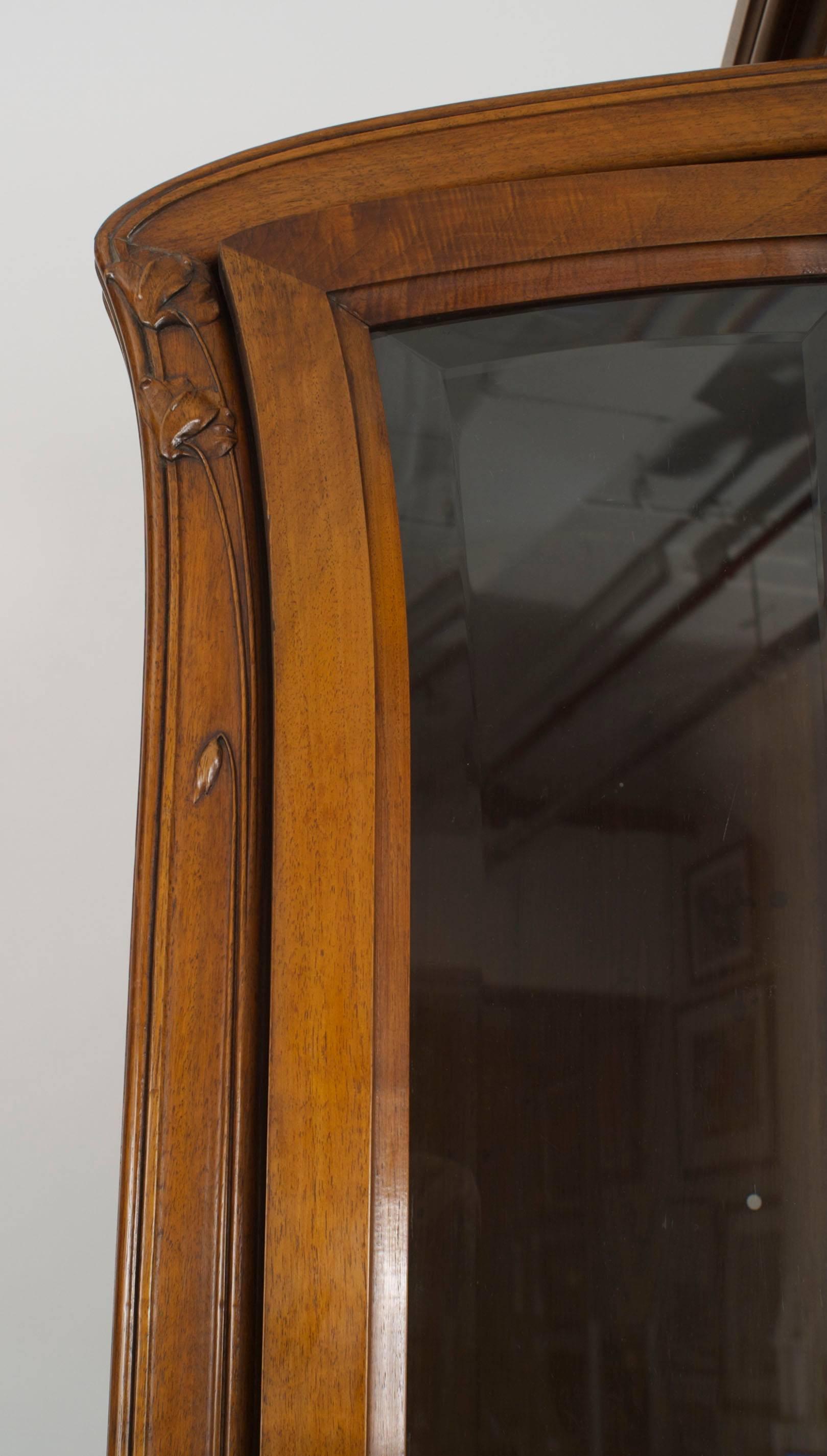 French Art Nouveau walnut display cabinet with 3 beveled glass doors and a floral carved center pediment and side corners.
