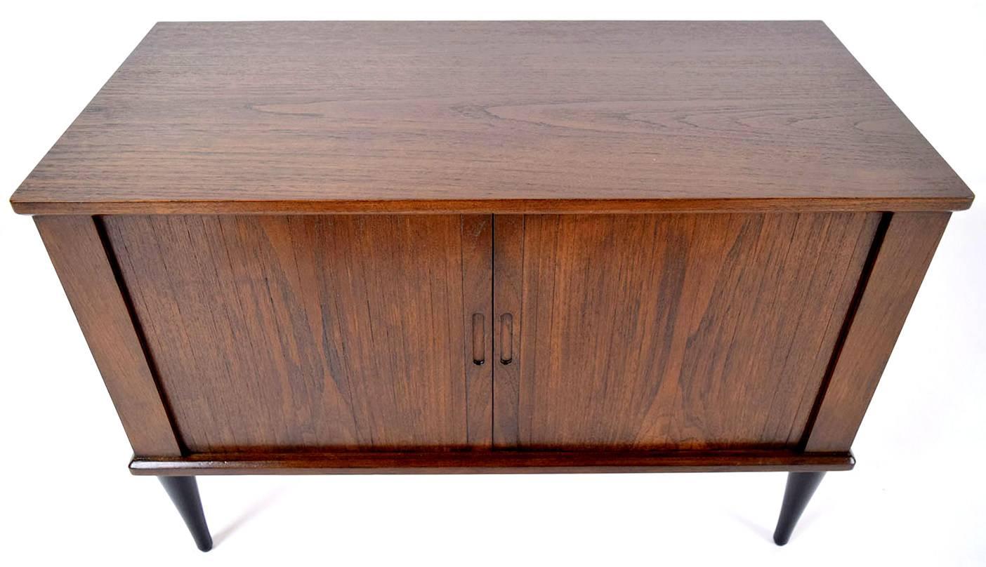 This 1960s Danish Mid-Century Modern style nightstand is made of teakwood finished in a rich walnut color stain. The Tambor cabinet doors open to reveal three compartments inside. There are two vertical compartments that flank a center compartment
