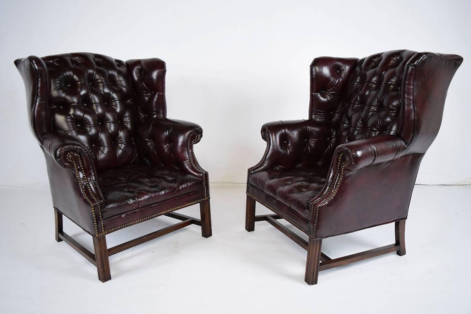 This 1950s vintage pair of Regency-style chesterfield wingback chairs feature their original leather upholstery. The chairs are elegantly adorned with tufted upholstery details on the seats, chair back, and arms. The edges feature nailhead trim
