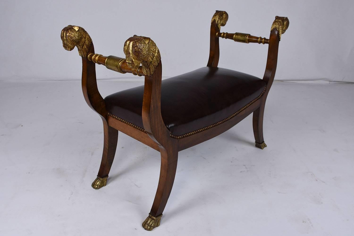 This 1900s antique French Empire-style bench features a frame made from mahogany wood in a beautiful rich stain finish. The frame is serpentine shaped with carved gilt details on the top of the handles of cat-esque figurine heads and finished with