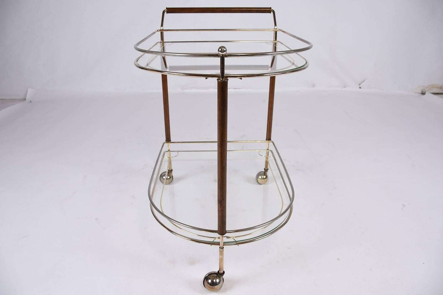 This 1960s Vintage Mid-Century Modern-style serving cart features a two-tier brass frame. Each tier is simply designed with a simple rail and glass bottom. The vertical poles and handle accent the brass base with teakwood finished a rich walnut
