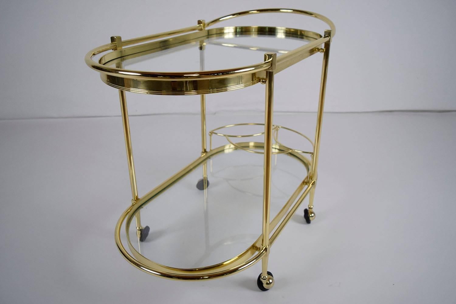 This 1980s vintage Italian Regency style oval tea cart features a brass frame. The brass frame features two levels. The top level features a glass shelf with a bar around the top with a handle to push the cart. The bottom shelf features a glass