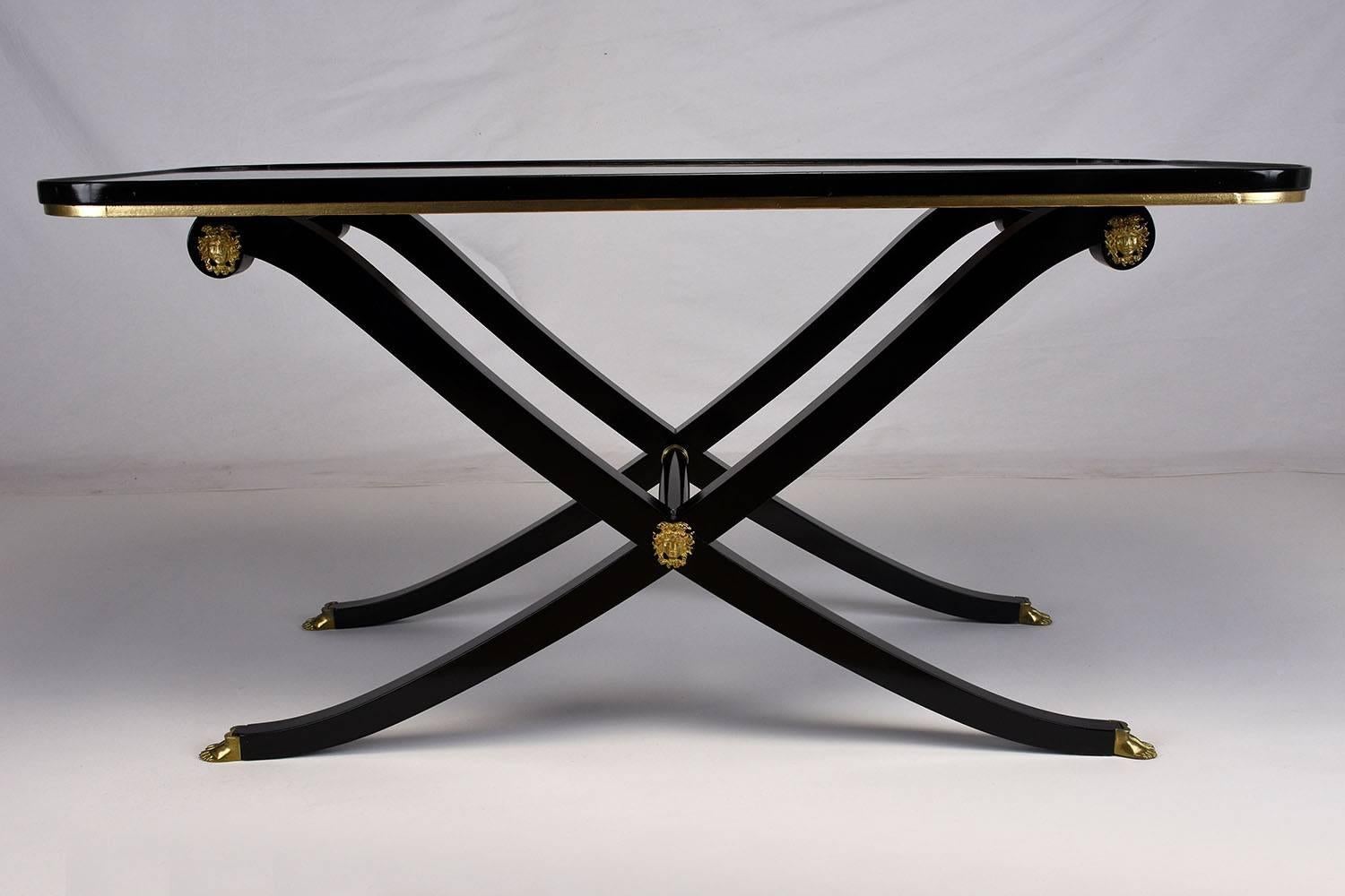 This 1950s vintage French Empire-style coffee table is made of mahogany wood that has been finished in a rich black color using an ebonizing technique. The wood top features a slight lip and is painted gold along the edge. The stretched X-shaped