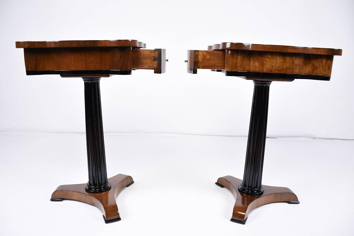 This stunning 1970's Vintage pair of Empire-style end tables are made of mahogany wood finished in a walnut color stain and a black stain on the pedestal legs. The undulating edge of the table top beautifully accents the square inlaid burlwood