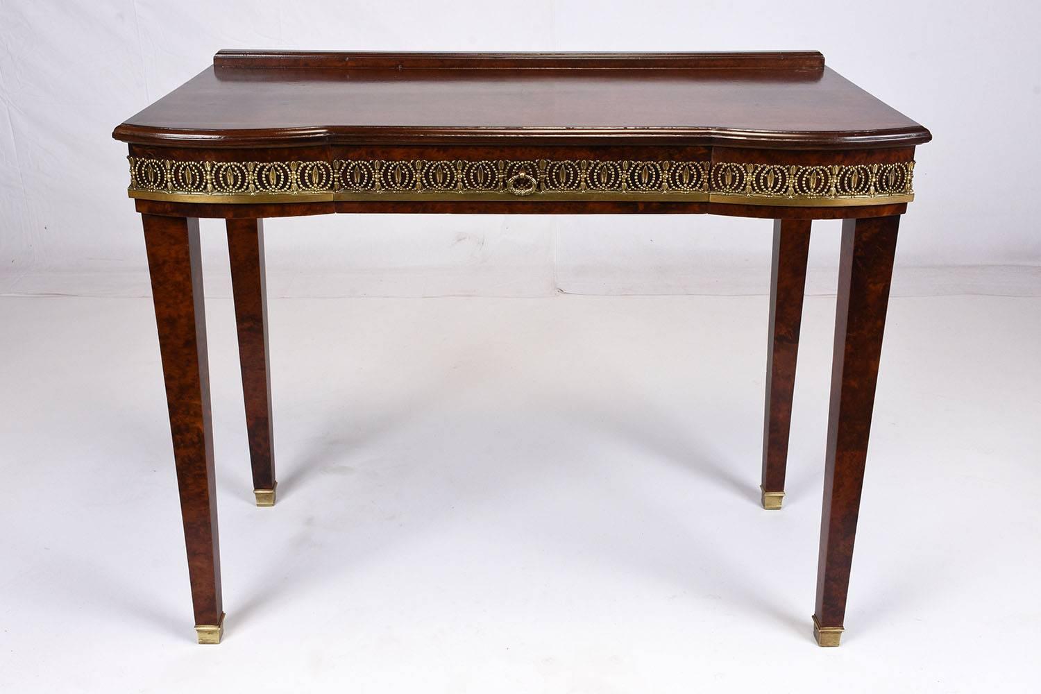 This 1900s Antique French Louis XVI style lady's writing table is made of solid mahogany wood and is covered in burl wood veneers in a rich dark wood tone lacquered finish. There is one centre drawer that opens with a decorative brass drawer pull.