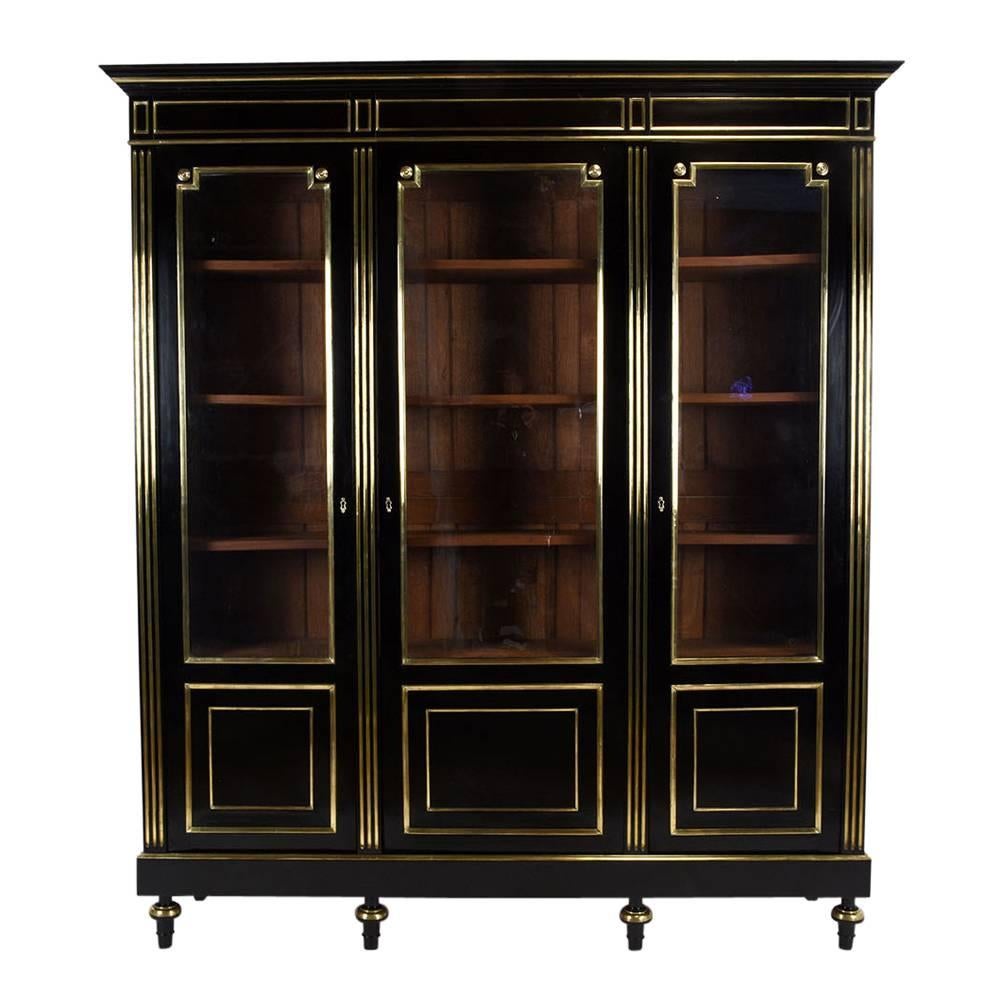 This 1890s French Louis XVI style bookcase is made of mahogany wood in a rich black color from an ebonized finish. Adorning the bookcase are brass moulding inserts and details on the front and sides. The three doors feature the original glass