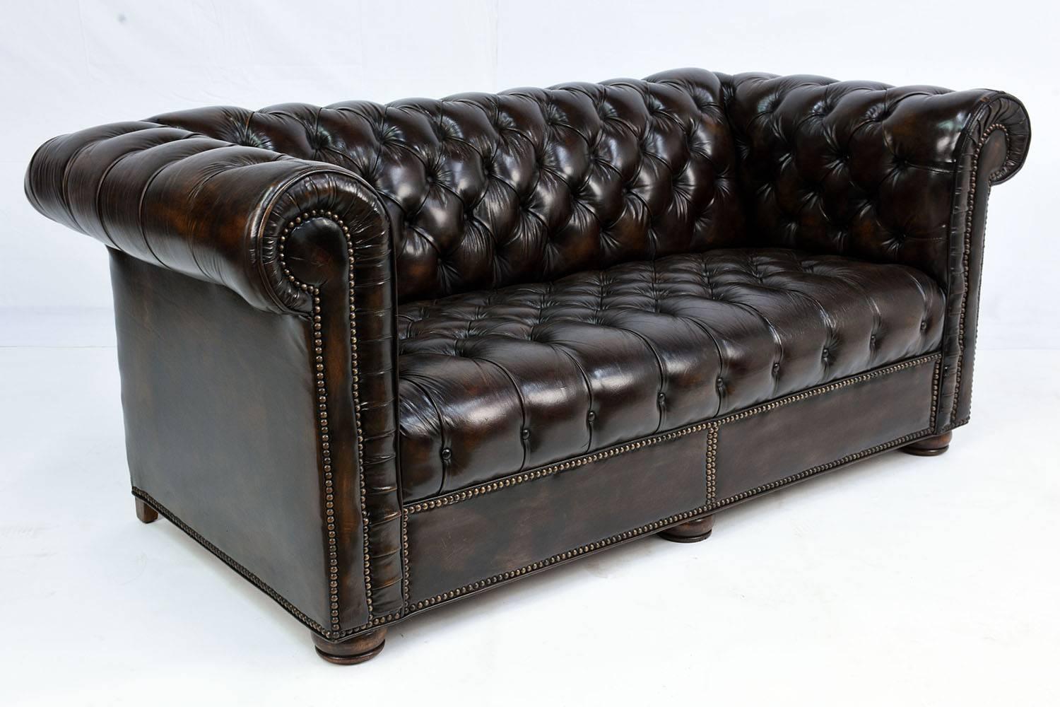 This 1970s vintage Chesterfield style love seat is upholstered in dark brown color leather with a patina finish. The seat, back and arms have elegant tufted details. The edges have brass nailhead decorations and the love seat is finished with round