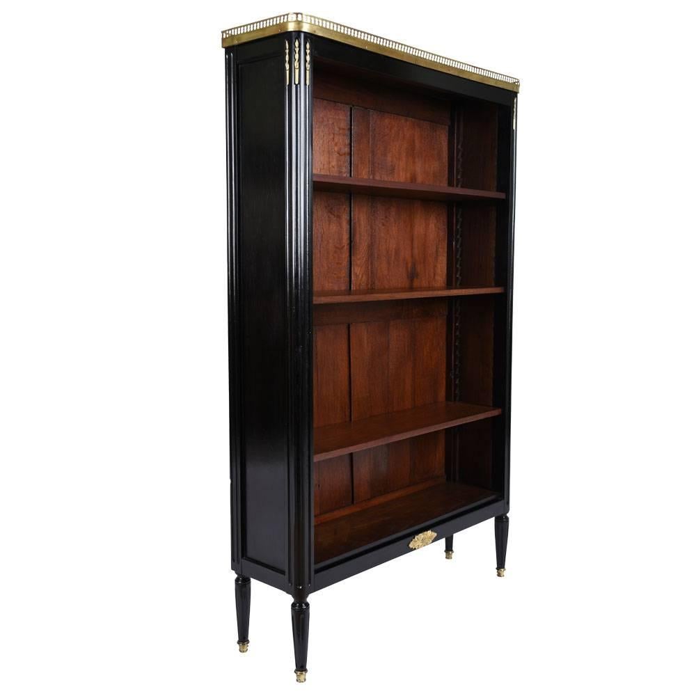This 1900s antique French Louis XVI-style bookshelf is made of mahogany wood that features a stunning ebonized finish. On top of the bookshelf there is a white colored marble top that is delicately bordered by a brass gallery with arched details.