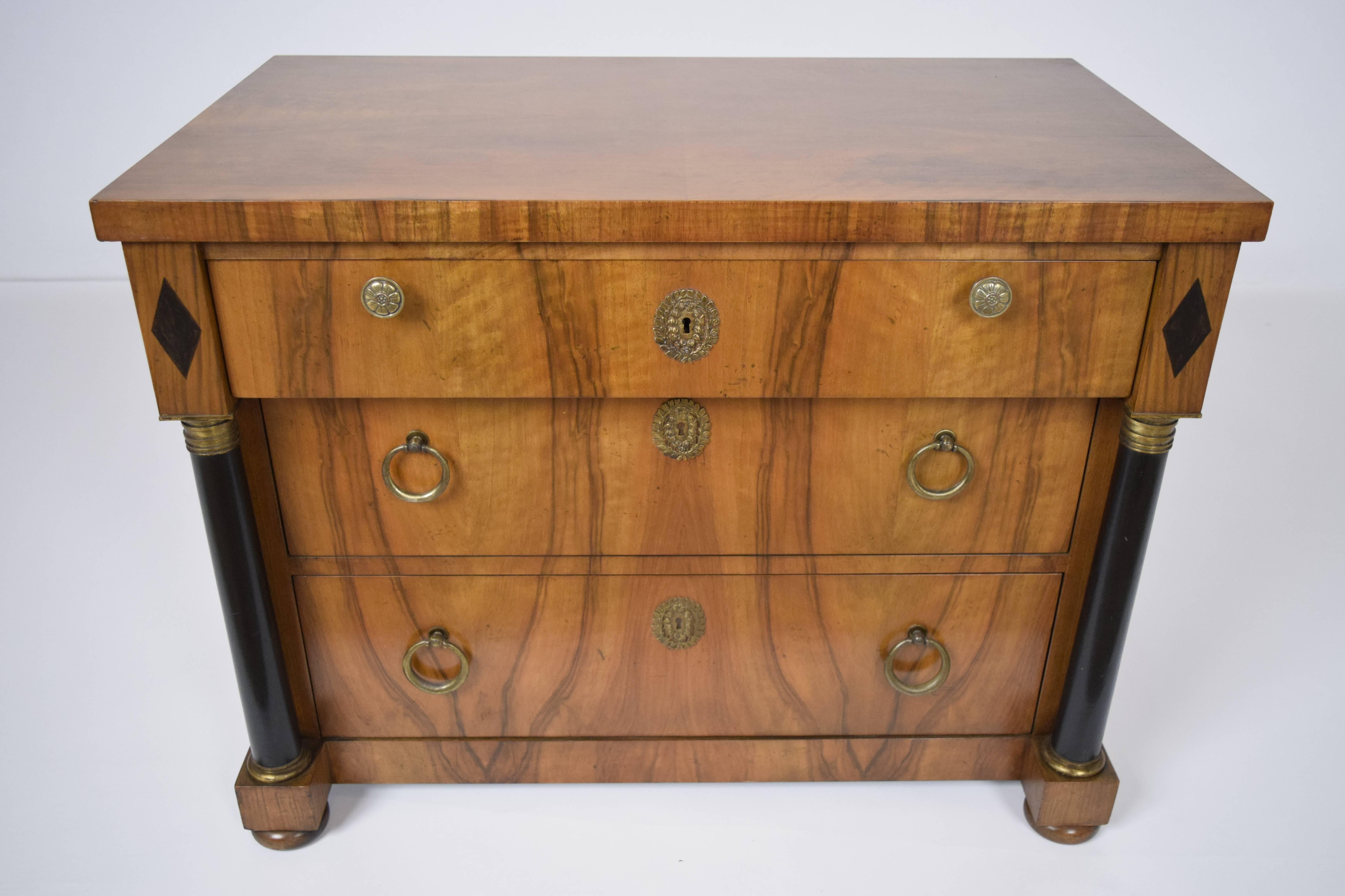 This 1960s vintage Empire-style chest of drawers is made by Baker. The chest is made of walnut wood that is finished in light walnut color stain. There is a beautiful wooden top with its wood grain visible. There are three drawers that are adorned