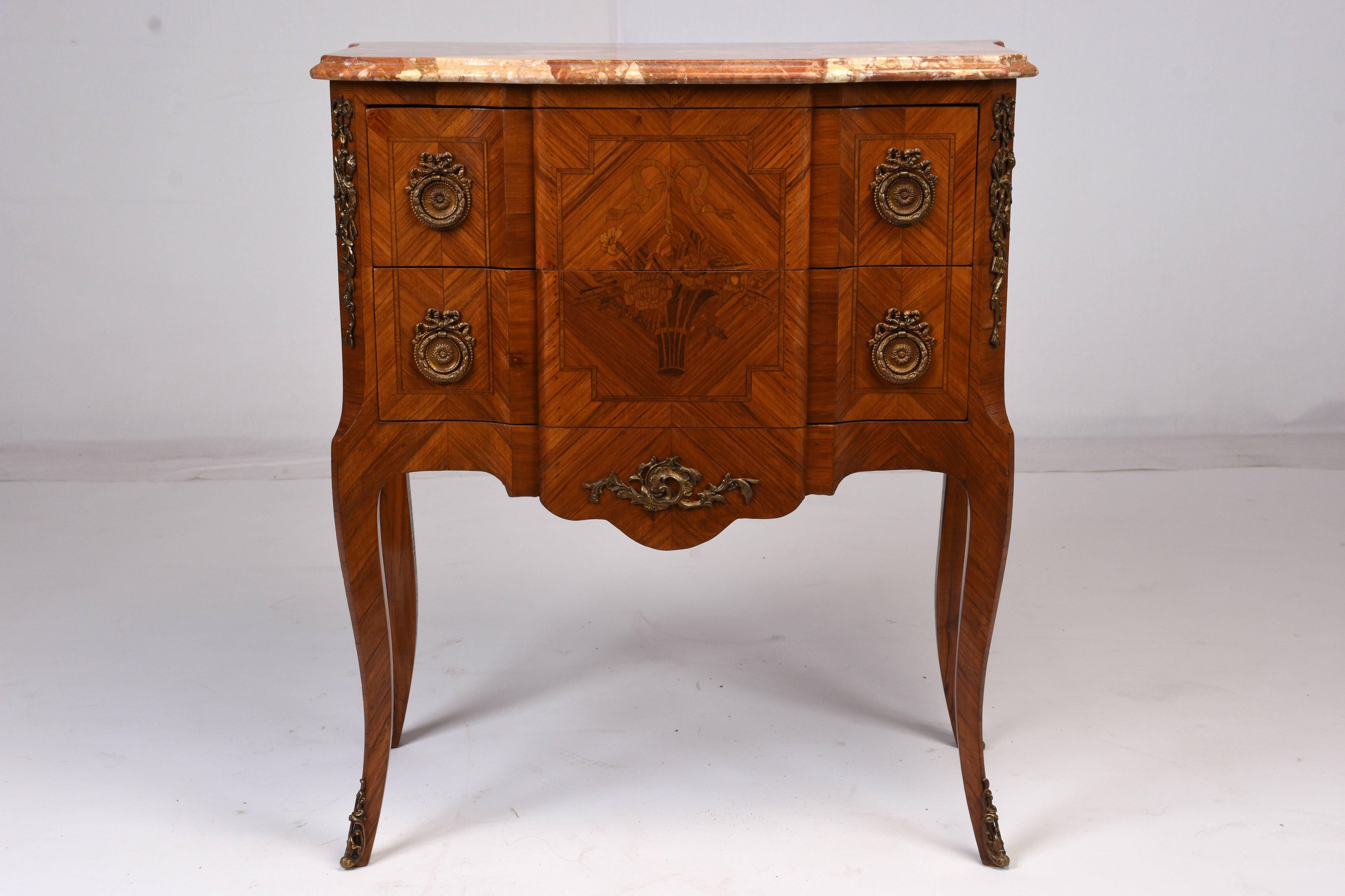 This 1950s antique French Louis XVI-style commode is made of wood adorned with stunning marquetry details and stained in a mahogany color. The front of the chest features geometric designs and a basket of flowers and a ribbon in the centre. The two