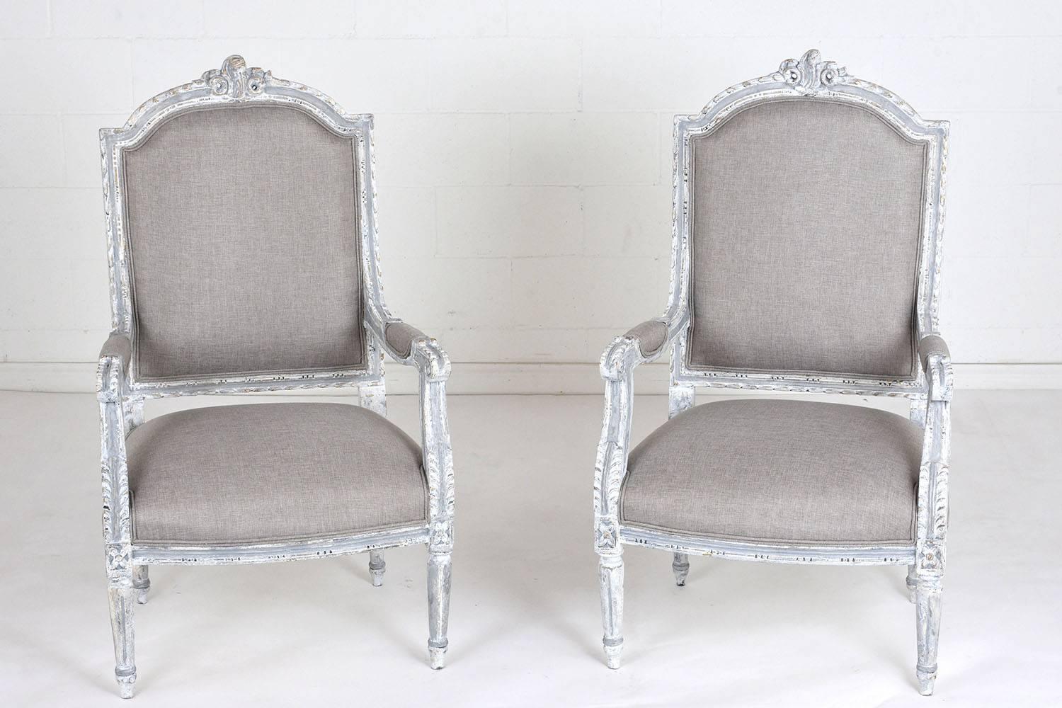 This pair of 1900s antique French Louis XVI-style armchairs feature wood frames painted in an off-white and grey color combination with a distressed finish. The frames are ornately adorned with carved details of acanthus leaves, garland mouldings,