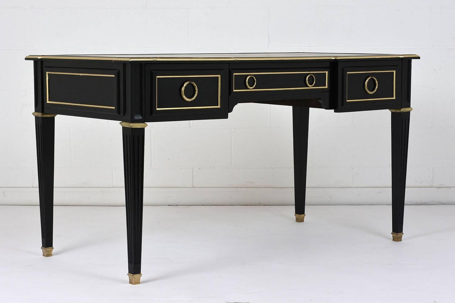 This 1900s antique French Louis XVI-style desk is made of mahogany wood with a deep ebonized and lacquered finish. The top features the original embossed leather insert with gilt details of acanthus leaves and scrolls. There are three drawers with
