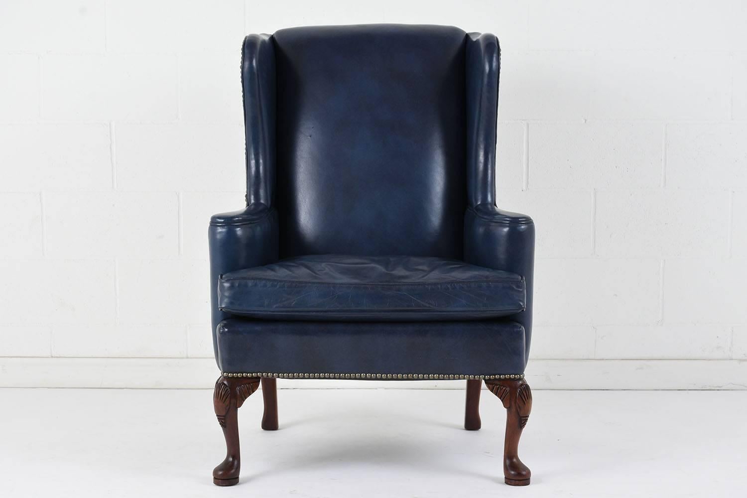 This 1970s Regency-style wing back chair features the original blue leather upholstery covering the majority of the chair. The seat has classic wing shaped sides and arm rests with scroll details extending outward. The comfortable seat cushion