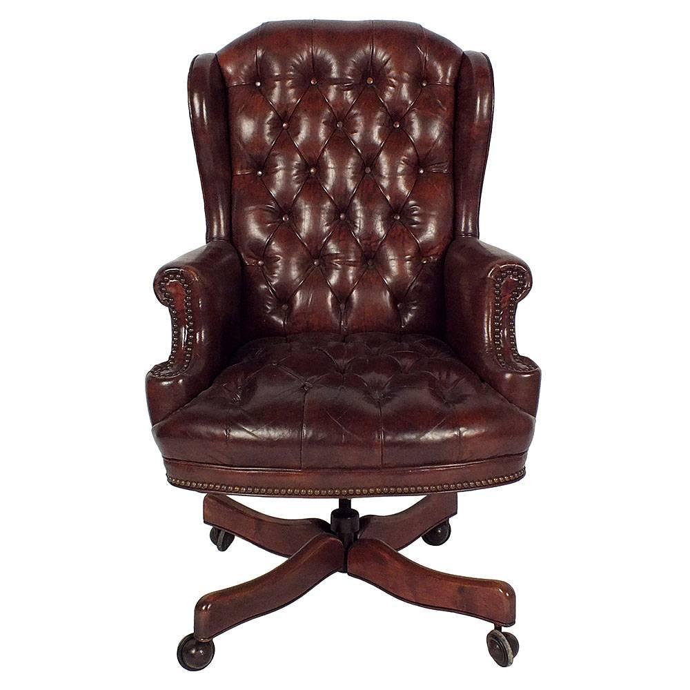 This 1960s Regency-style office chair features leather upholstery and a wood base. The leather upholstery has tufted details on the seat and back and is accented by nail head trim. The wingback chair is raised by a pedestal base with wood legs and