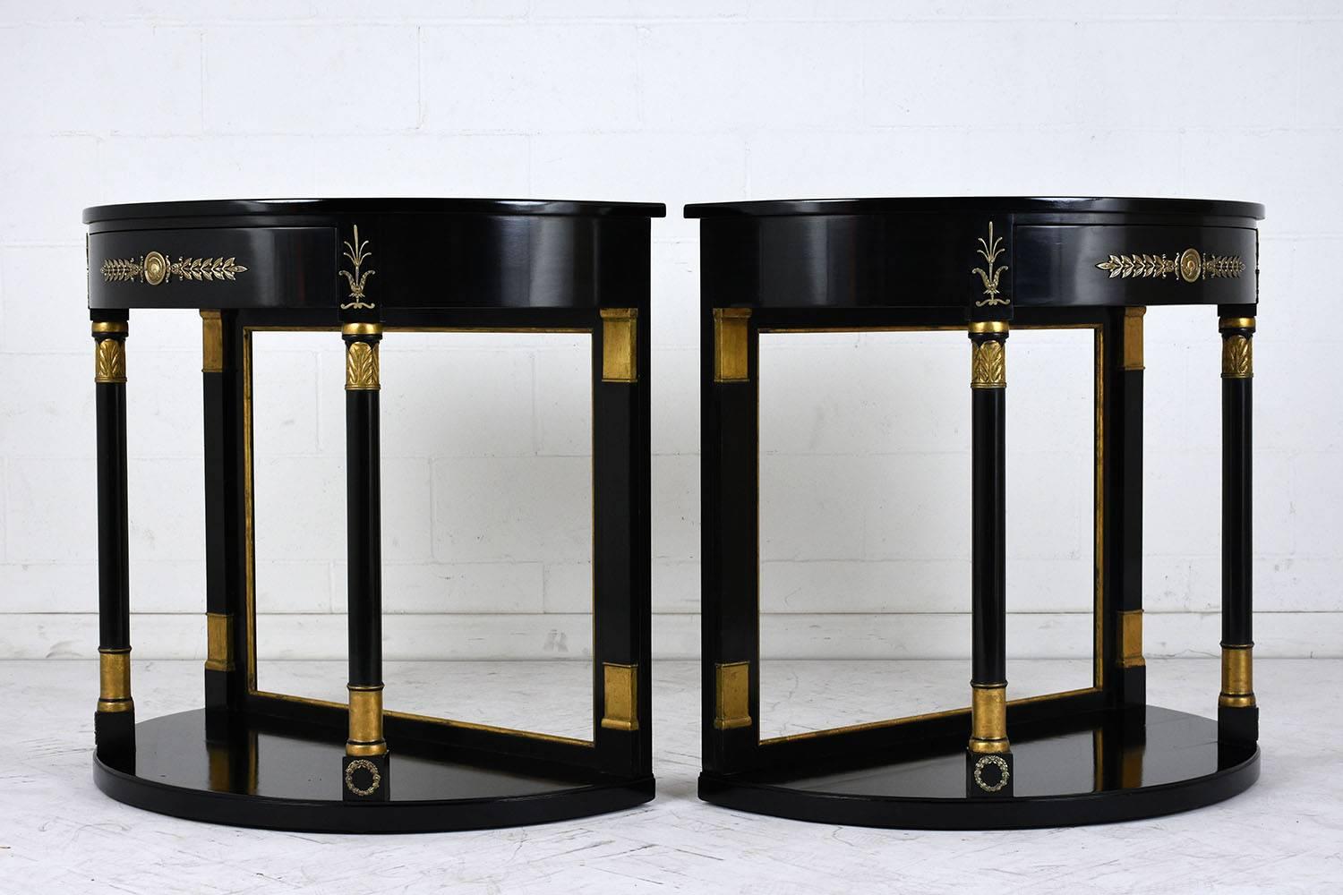 This pair of 1950s Empire-style demi-lune console tables are made of wood that has been ebonized with a lacquered finish. The console tables have a single drawer that opens from the bottom edge and is adorned with ornate brass accents. Below the