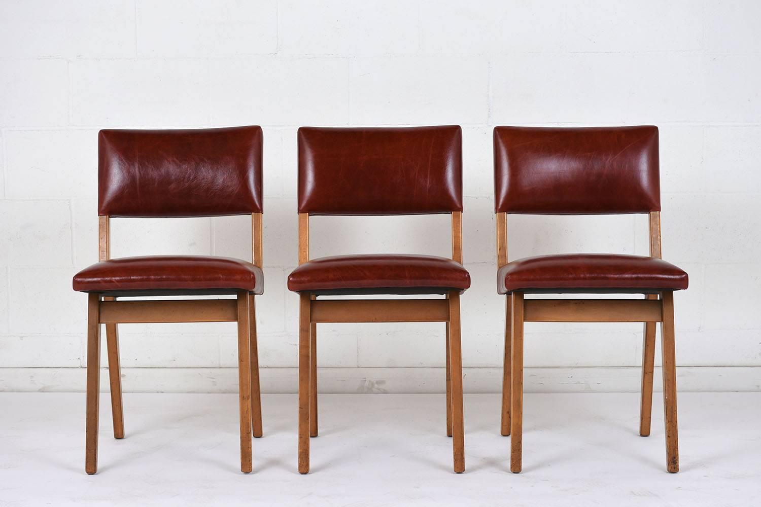 This set of six Mid-Century Modern-style dining chairs have a wood frame finished in a light walnut color. The seats and back have recently been professionally upholstered in brick red leather with brass nailhead details. This set of dining chairs