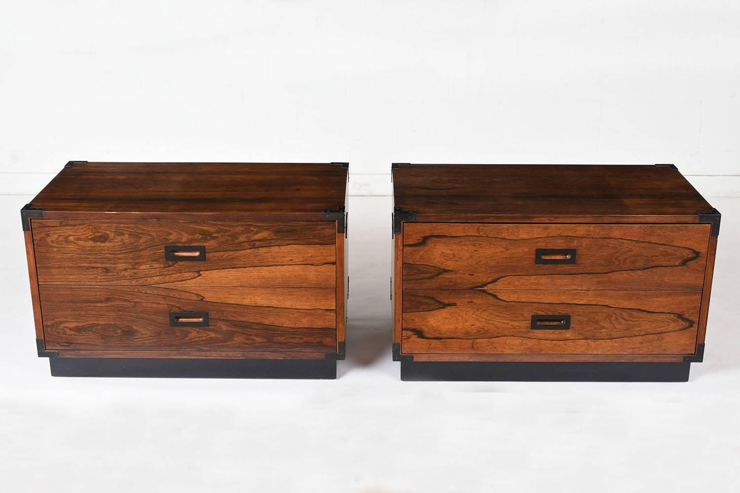 This pair of 1960s Campaign-style chest of drawers is made of rosewood stained a rich rosewood color with a lacquered finish. The chests have black metal accents on the corners and around the carved drawer pulls. Each chest has two drawers with