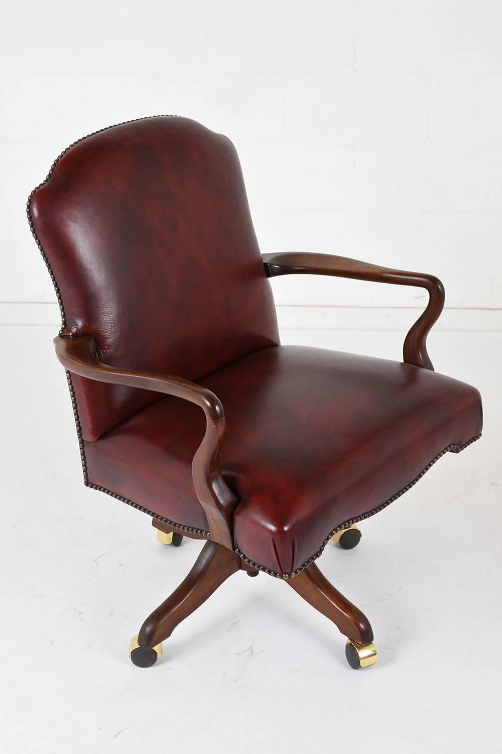 This Regency-Style Swivel Office Chair is upholstered in a burgundy color leather that is accented by brass nailhead trim along the edges. The arms and base are made of mahogany wood stained in rich mahogany color, and the chair features a mechanism