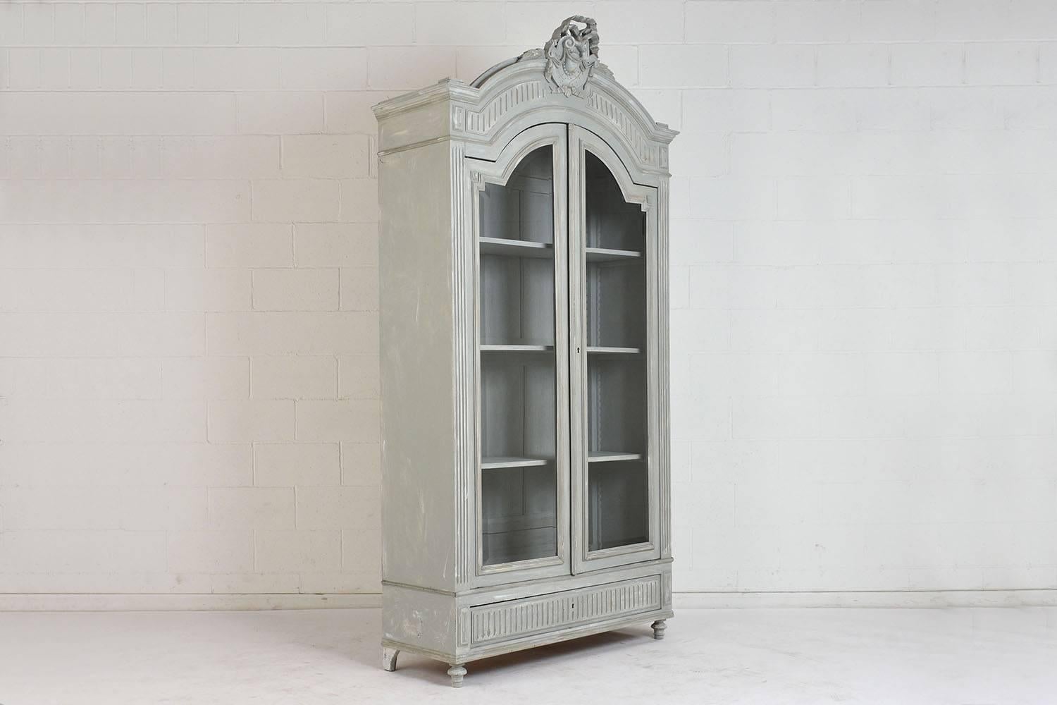 This 1900s French Louis XVI-style bookcase is made of walnut wood painted in a pale gray and off-white color combination with a distressed finish. The top of the bookcase features a carved crest of ribbons, garlands, leaves, and scrolls. The front
