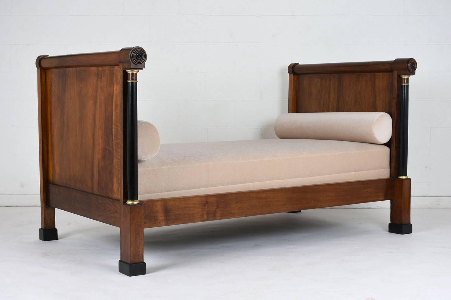 This 1830's French Empire-style daybed features a completely restored walnut wood frame stained a rich walnut color with black and gilt accents. The comfortable new mattress cushion is upholstered in a beige mohair velvet fabric with two matching