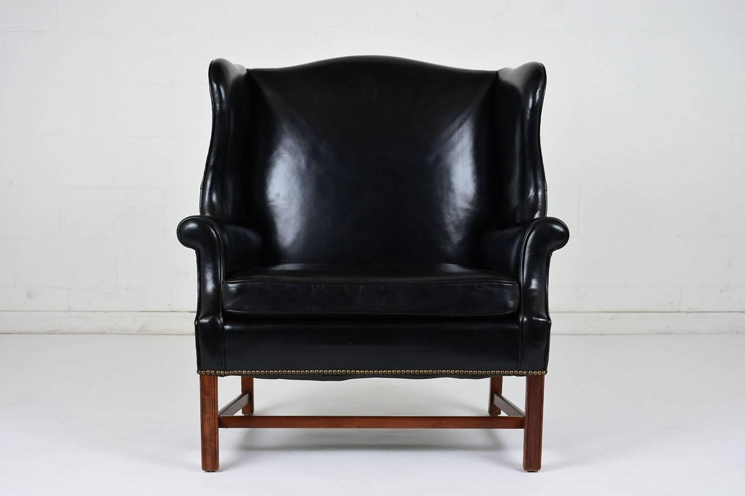 This 1930's English Regency-style wingback chair features an very wide seat and is completely upholstered in the original black color leather with single piping trim details. The curved side wings and top edge flow into the scroll arm rests. The