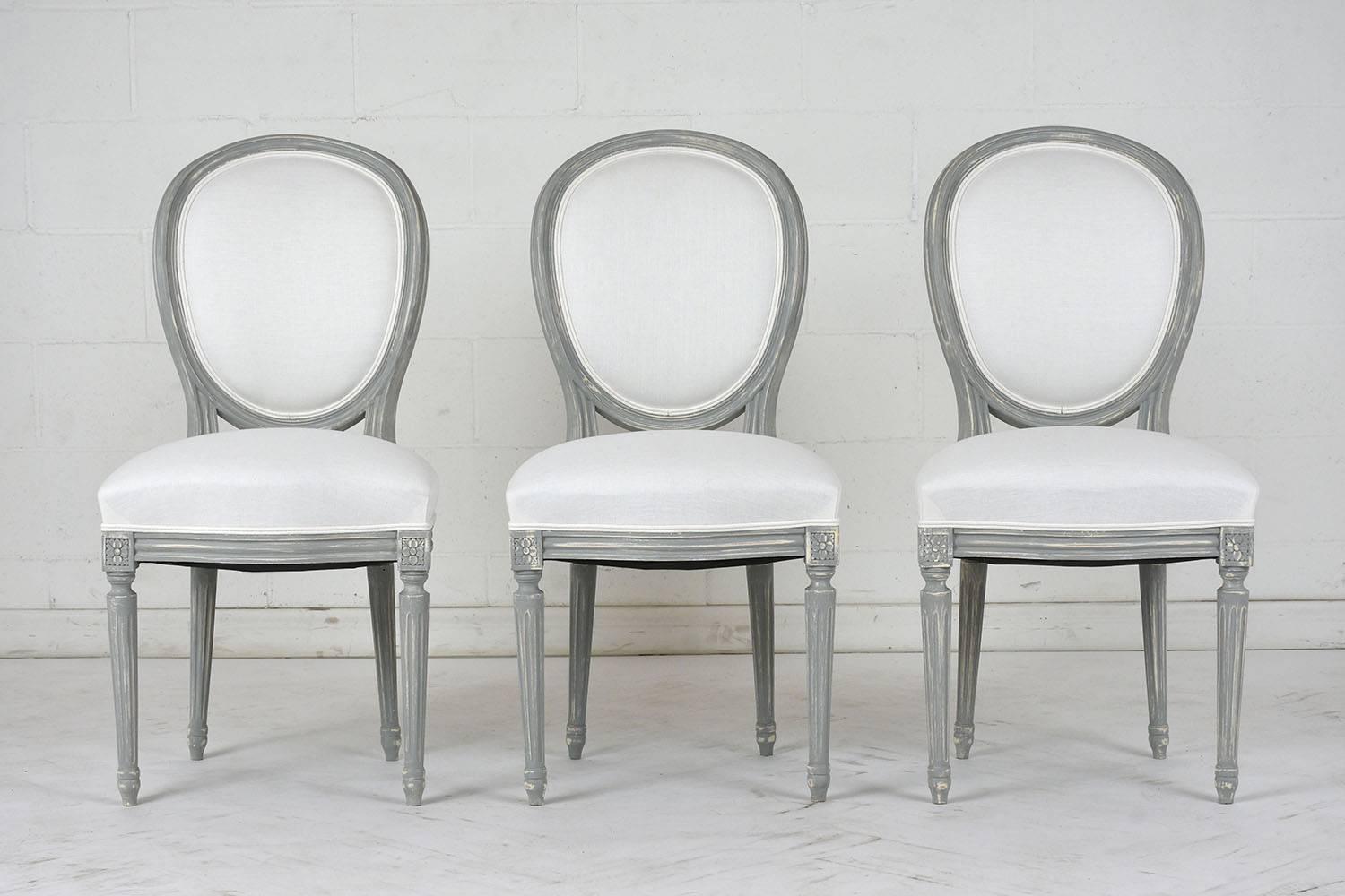 This set of six 1910s French Louis XVI-style dining chairs have a curved oval back and carved walnut wood frame finished in a pale grey and off-white color combination with a distressed finish. The frame has fluting and rosette carved details