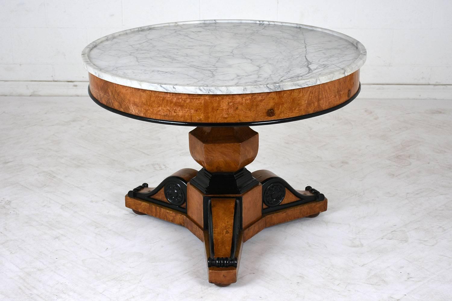 This 1850s French Empire-style round center table is made of wood covered in burl veneers stained a rich golden color with a lacquered finish. The pedestal base features a bucket shape with ornately carved legs. The legs have black accents on the