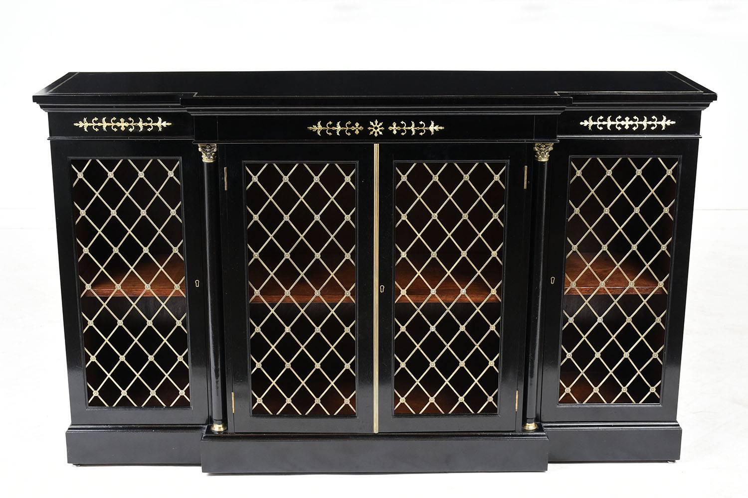 This 1910s Regency-style bookcase is made of mahogany with an ebonized and lacquered finish. The wood top features a brass boarder accent. The bookcase has carved columns flanking the jutted out center section and brass accents on the columns and