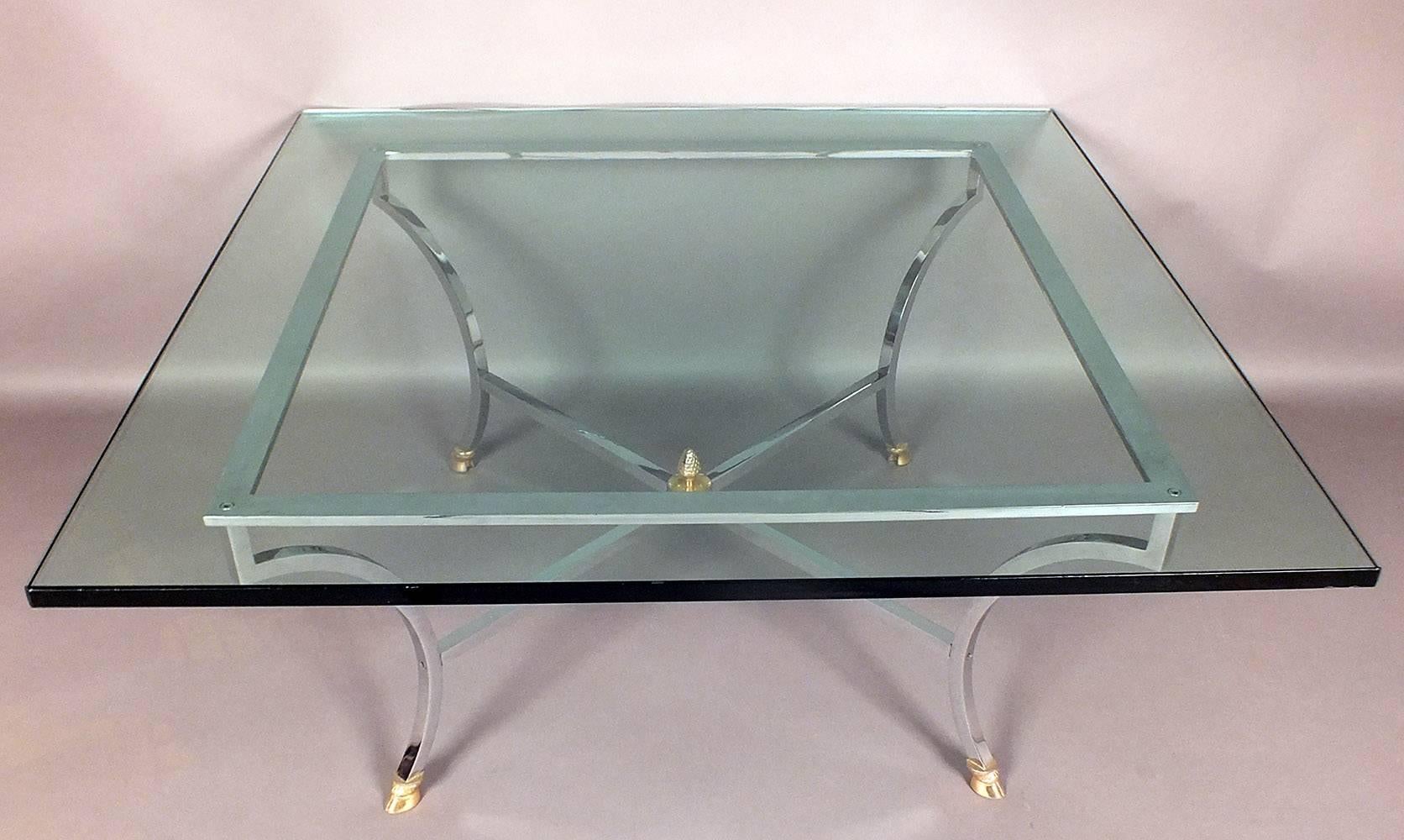 This 1970's Vintage Hollywood Regency-style coffee table features a square glass top that is 3/4 inches thick with a flat polish finish. Holding up the glass is a sleek square brass frame in a chrome color with X-shaped stretched legs and a finial