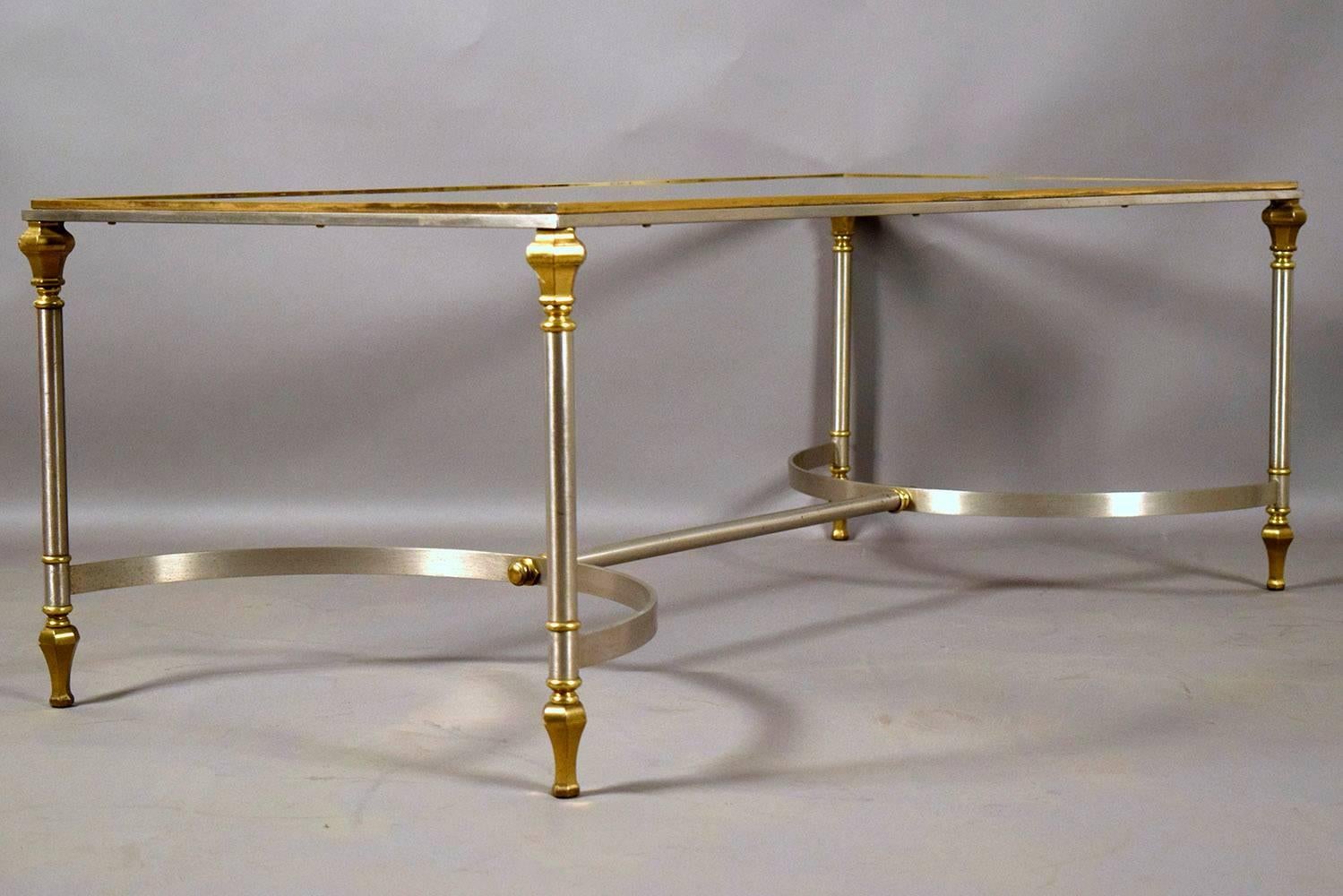 This Hollywood Regency-style Maison Jansen-style coffee table features an elegant brass and chrome frame. The legs of the coffee table feature vertical fluting and decorative capitals and feet. The legs are strengthened by half circle stretched