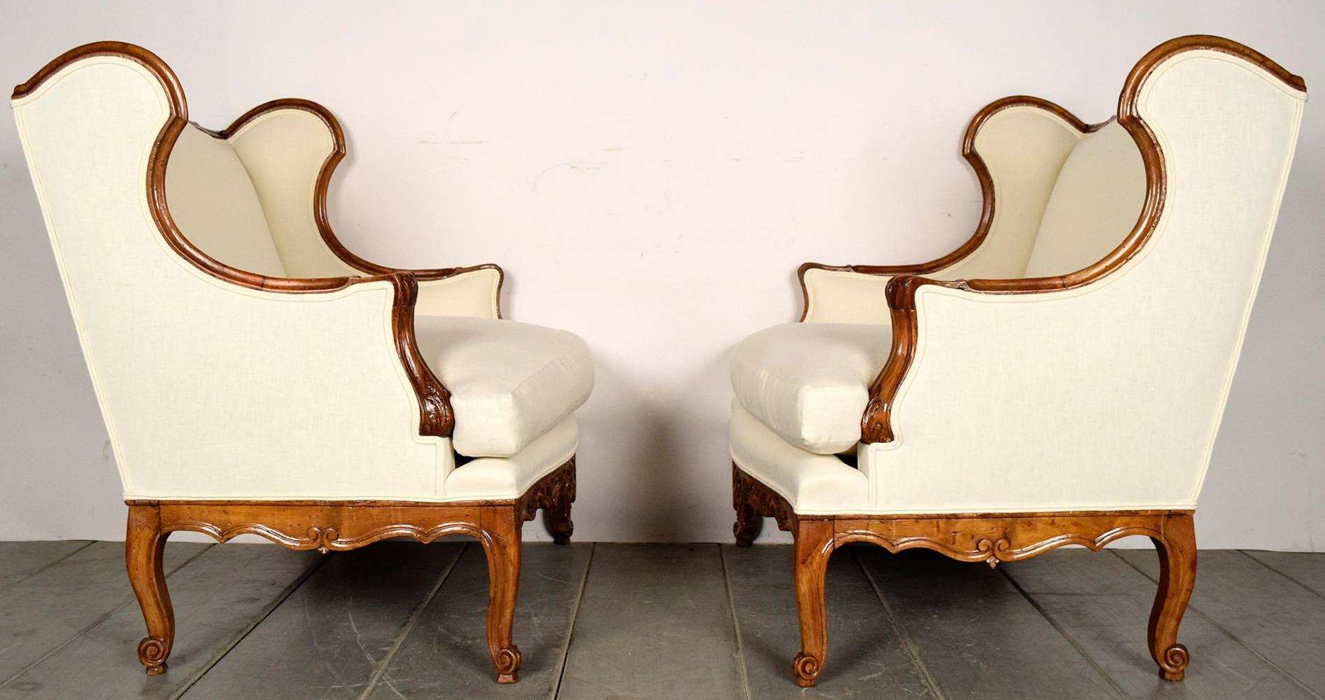 Pair of 19th century wing chairs in Louis XVI-style. Solid wood frames, in its original walnut color finish. Carvings all throughout the frame, with a center crest on the center bottom. Newly upholstered in a ivory linen fabric, with double piping
