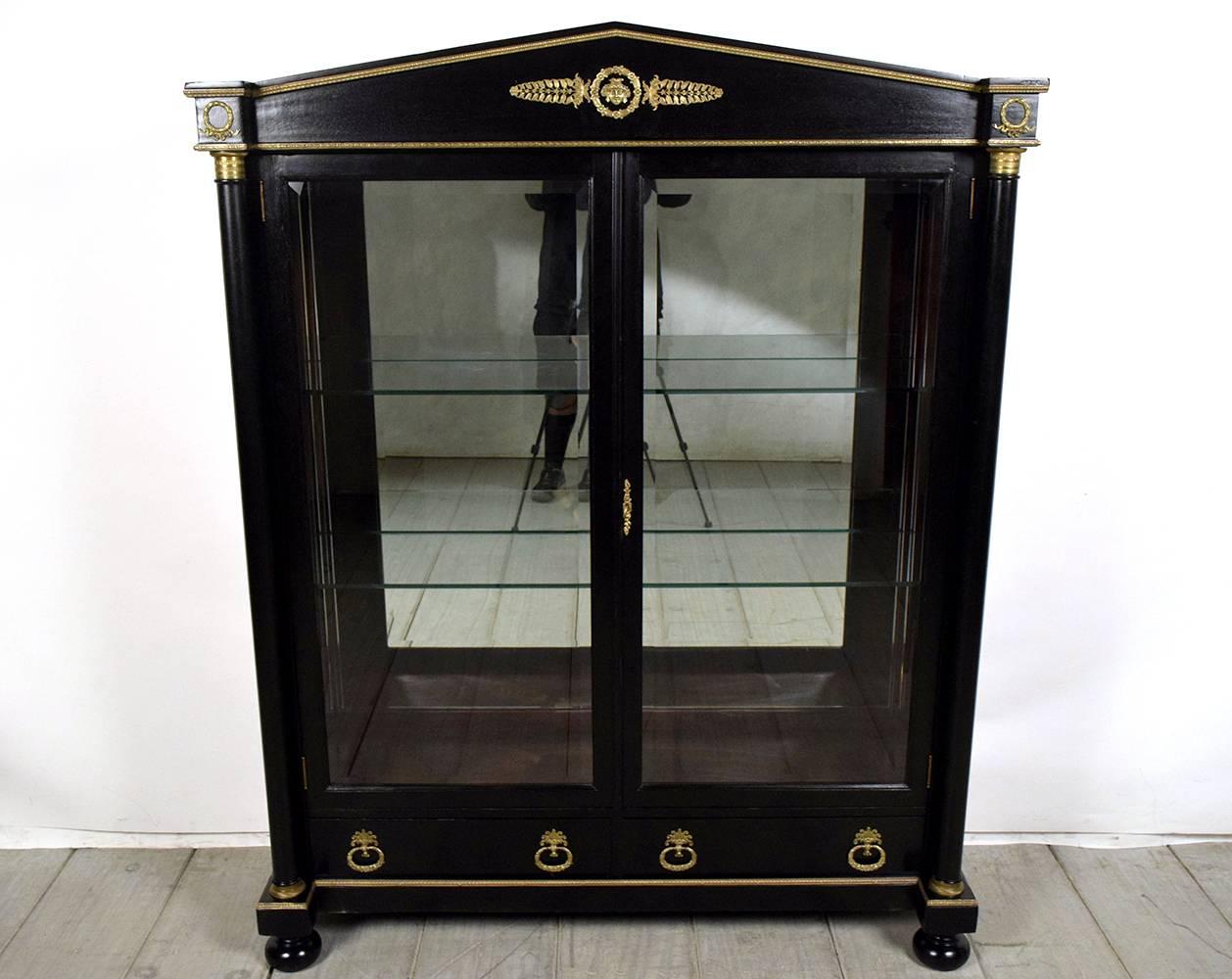 This 19th Century French Empire-style two door display Cabinet is recently finish in a black color with a lacquer finish. There are bronze ormolu decorations through out the facade of the frame. At the center of the top frame there is a bronze face