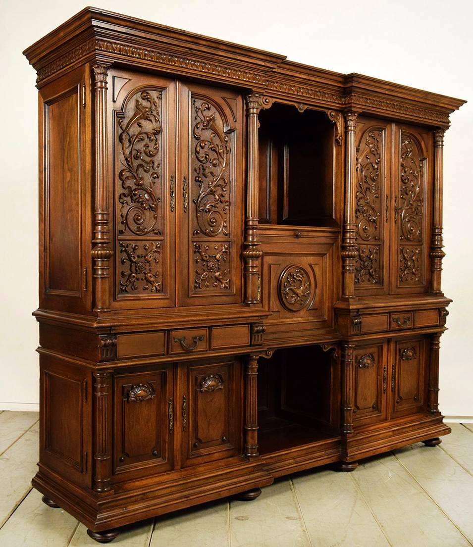 This 19th century renaissance grand cabinet features intricate decorations heavily carved into the solid walnut wood. There are four doors on top with engraved floral and scroll motif details on all panels. In the centre there is an open shelf which