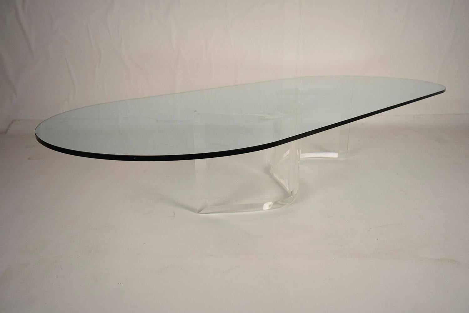 This 1970's Mid-Century Modern coffee table features an oval glass top with a Lucite S-shaped base. The glass top and clear lucite base allow for this coffee table to blend in with its surroundings while still drawing attention to itself. This